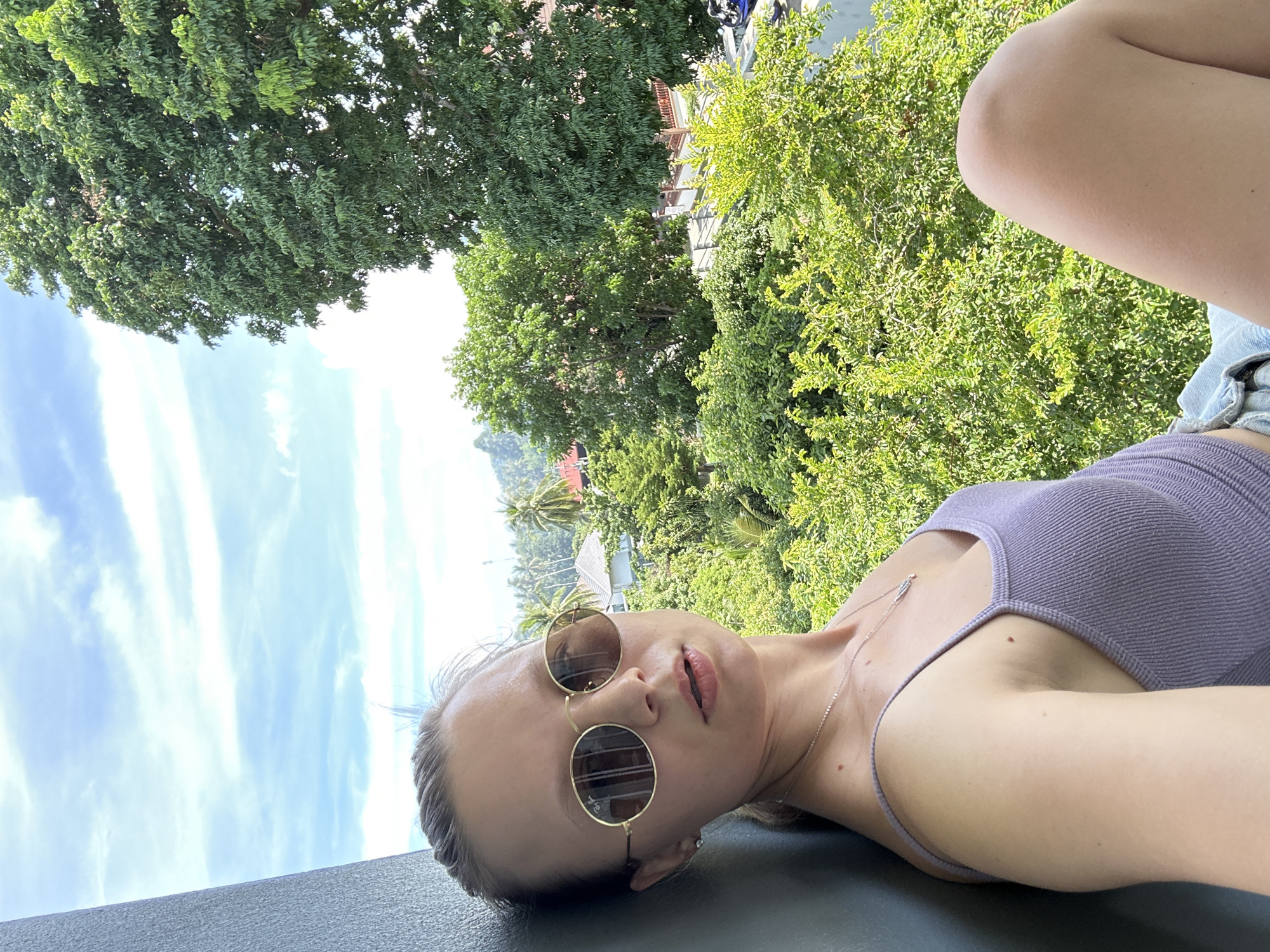 In Baan Hua Thanon, Thailand, a young woman with blonde hair pulled back and wearing round, gold-rimmed sunglasses and a lilac ribbed tank top, poses for a selfie on a balcony. She leans against a dark gray wall, her left arm visible. The background reveals a lush, green landscape with dense foliage and trees, indicative of a tropical setting. In the distance, houses with red tiled roofs and palm trees can be seen, suggesting a residential area. The sky is partly cloudy with a bright blue expanse. The overall scene captures a relaxed, outdoor moment in a vibrant, green environment.