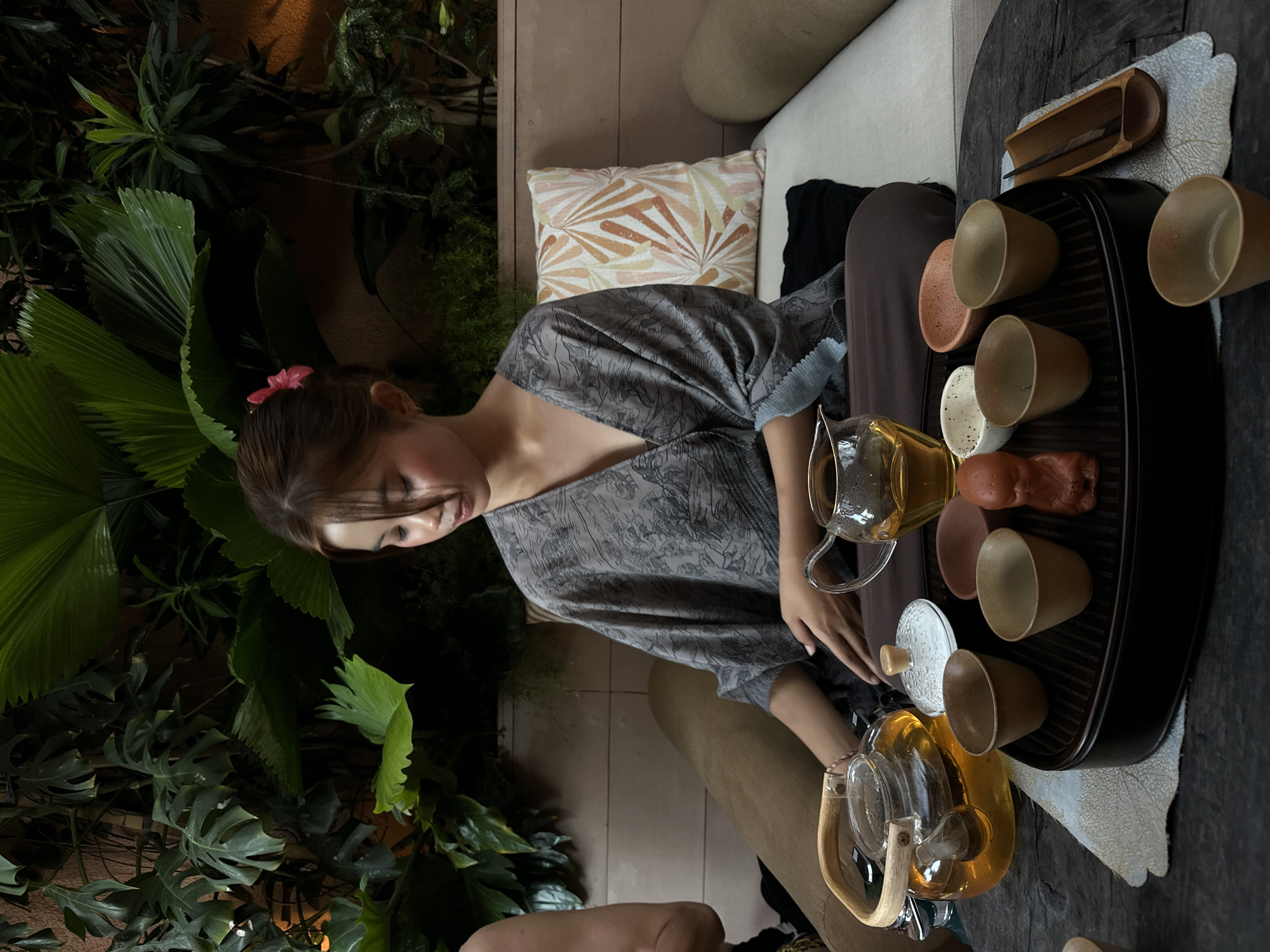 A woman is sitting at a table preparing tea in Pecatu, Indonesia. The woman has dark hair and is wearing a gray dress with floral prints and a small pink flower in her hair. She is looking down at the table, where she is arranging teacups and two glass teapots, one with a lid. One of the teapots is filled with a yellow liquid. The teacups are arranged on a black tray, along with a small wooden container. A decorative pillow with a leaf pattern is behind her, on what appears to be a light beige sofa. Lush green plants surround the area.