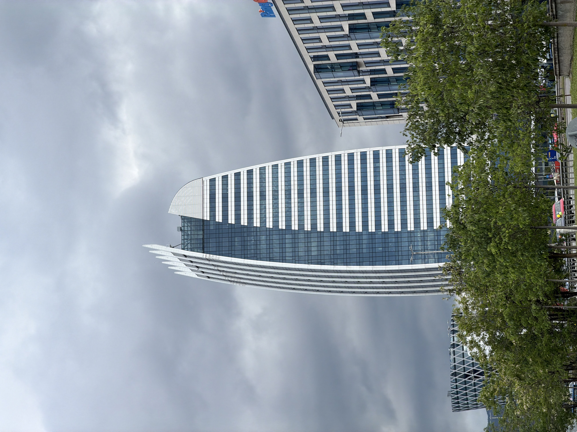 The image shows a view of several buildings in Sofia, Bulgaria, taken on an overcast day. The primary focus is on the Capital Fort office building, which is a modern, curved structure with a glass and white panel facade. In the foreground, there are trees partially obscuring the view along the street. Another tall, rectangular building with a grid-like exterior is visible above and behind Capital Fort. A third building with a crosshatched design is also partially visible in the lower-right corner of the image. The sky is filled with dark, grey clouds, suggesting an impending storm. Some street furniture, including possible traffic lights and poles, can be seen through the foliage. The image appears to have been taken from ground level.