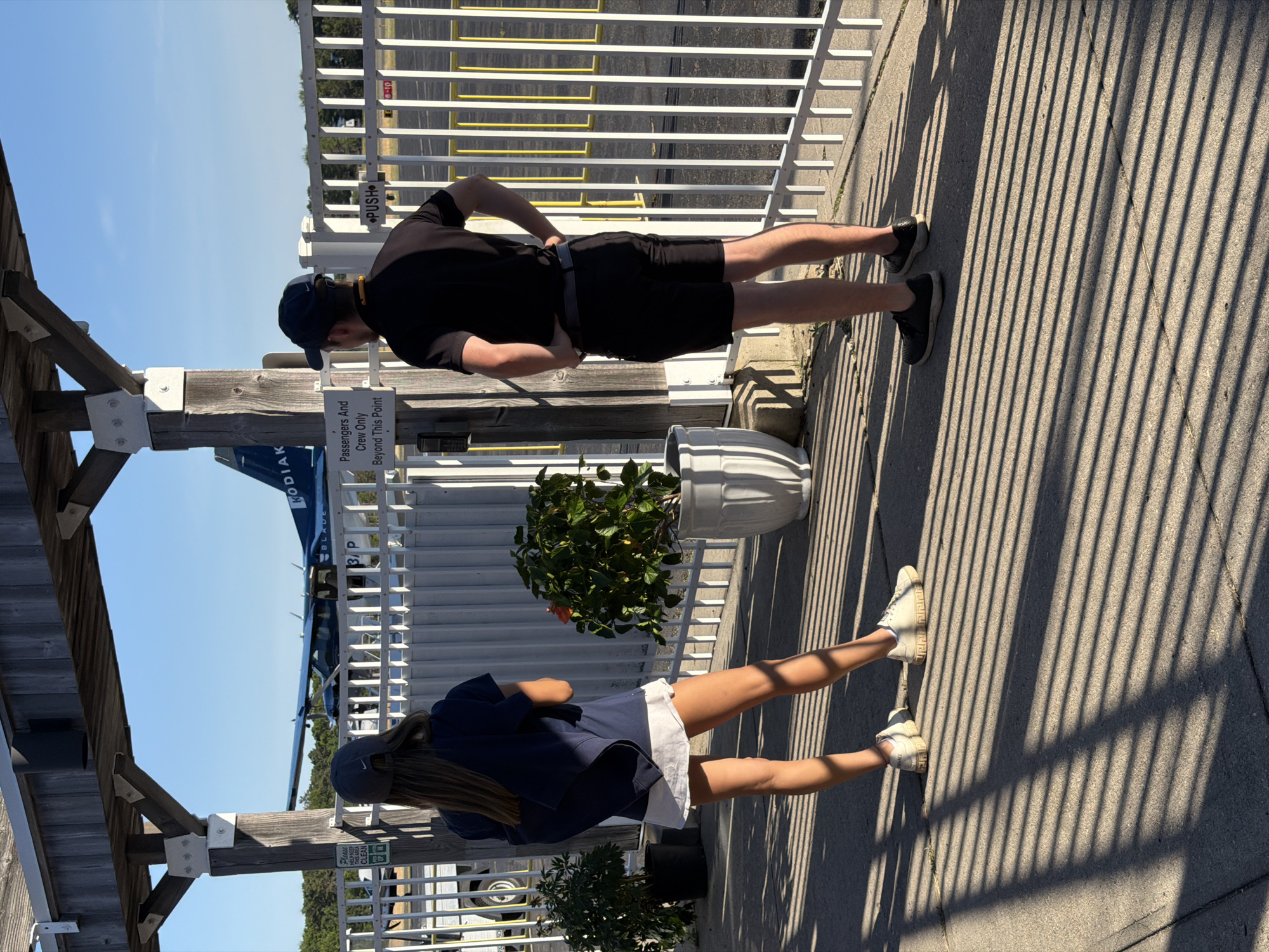 In the Town of East Hampton, United States of America, a young woman and a young man stand on a paved walkway outside a small airport. The woman, facing away from the camera, wears a navy blue t-shirt, a white skirt, a navy blue baseball cap, and white sneakers. She has a navy blue backpack slung over her shoulders. The man, also facing away, is dressed in a black short-sleeved shirt, black shorts, a black baseball cap, and black sneakers. Both individuals appear to be waiting.
Behind them, a white fence runs along the perimeter of the airport tarmac. A small propeller airplane with "KODIAK" visible on its tail is parked on the tarmac. The environment is sunny with distinct shadows cast by the fence on the ground. Two potted plants, one with green foliage and the other with a red flower, are placed near the fence. A sign on the fence reads "Passengers And Crew Only Beyond This Point." Another sign on the fence further down reads "PUSH." The scene suggests a departure or arrival at a small regional airport on a clear day.