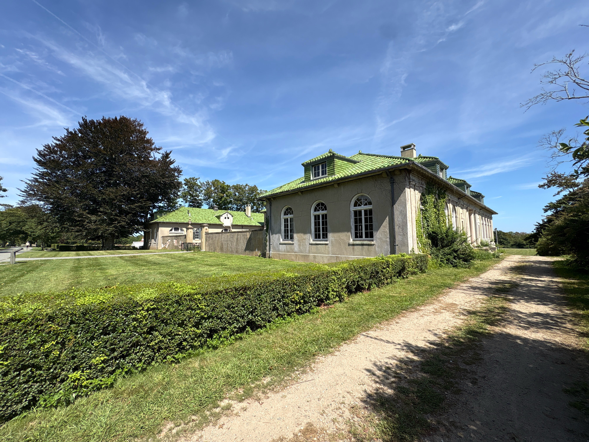 The image shows a large building with a green tiled roof, likely a mansion or estate, situated on a grassy lawn with mature trees. A dirt path runs alongside the building, bordered by a neatly trimmed hedge. The sky is a vibrant blue with scattered white clouds. In the background, another smaller building with a similar green roof is visible. The overall impression is one of a well-maintained, historic property in a rural or suburban setting. There are no people or animals visible in the immediate vicinity.