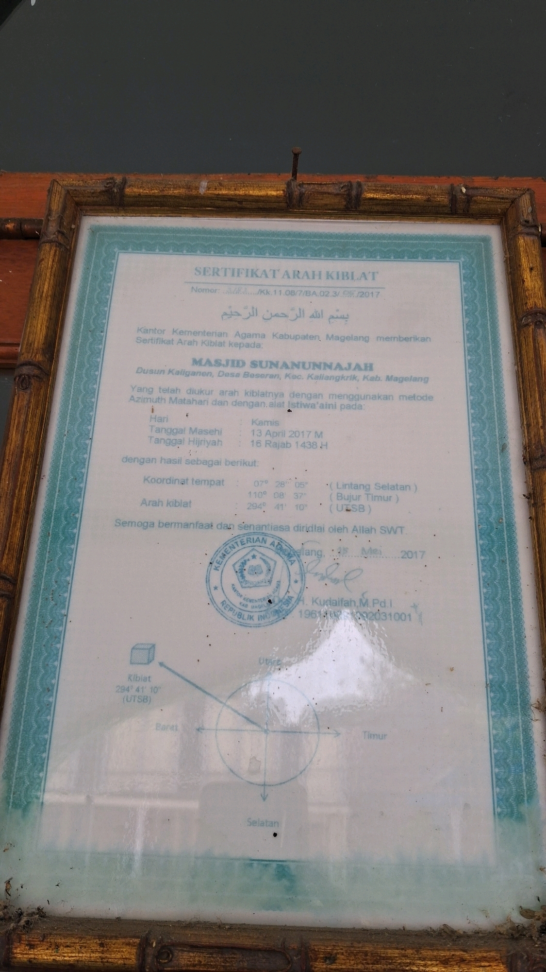 The image shows a framed certificate for the direction of the Qibla. The certificate is from the Ministry of Religious Affairs of Magelang Regency and is issued to the Masijd Sunanunnajah in Dusun Kaliganen, Desa Beseran, Kecamatan KallangKrik, Kabupaten Magelang. The certificate states that the Qibla direction was measured using the Sun's azimuth and the "Istiwa'aini" method. The coordinates for the location are given as 07° 28' 05" (Lintang Selatan) and 110° 08' 37" (Bujur Timur). The Qibla direction is specified as 294° 41' 10" (Utara). The certificate is dated 13 April 2017 M / 16 Rajab 1438 H. It is signed by H. Kudaifah, M.Pd.I. The bottom part of the certificate includes a compass rose indicating North, South, East, and West. The frame is made of wood and appears to be bamboo-like in texture. The certificate is behind glass and shows some reflections. The text on the certificate is in Indonesian. There is no visible indication of the city of Giriwarno, Indonesia, in the image.