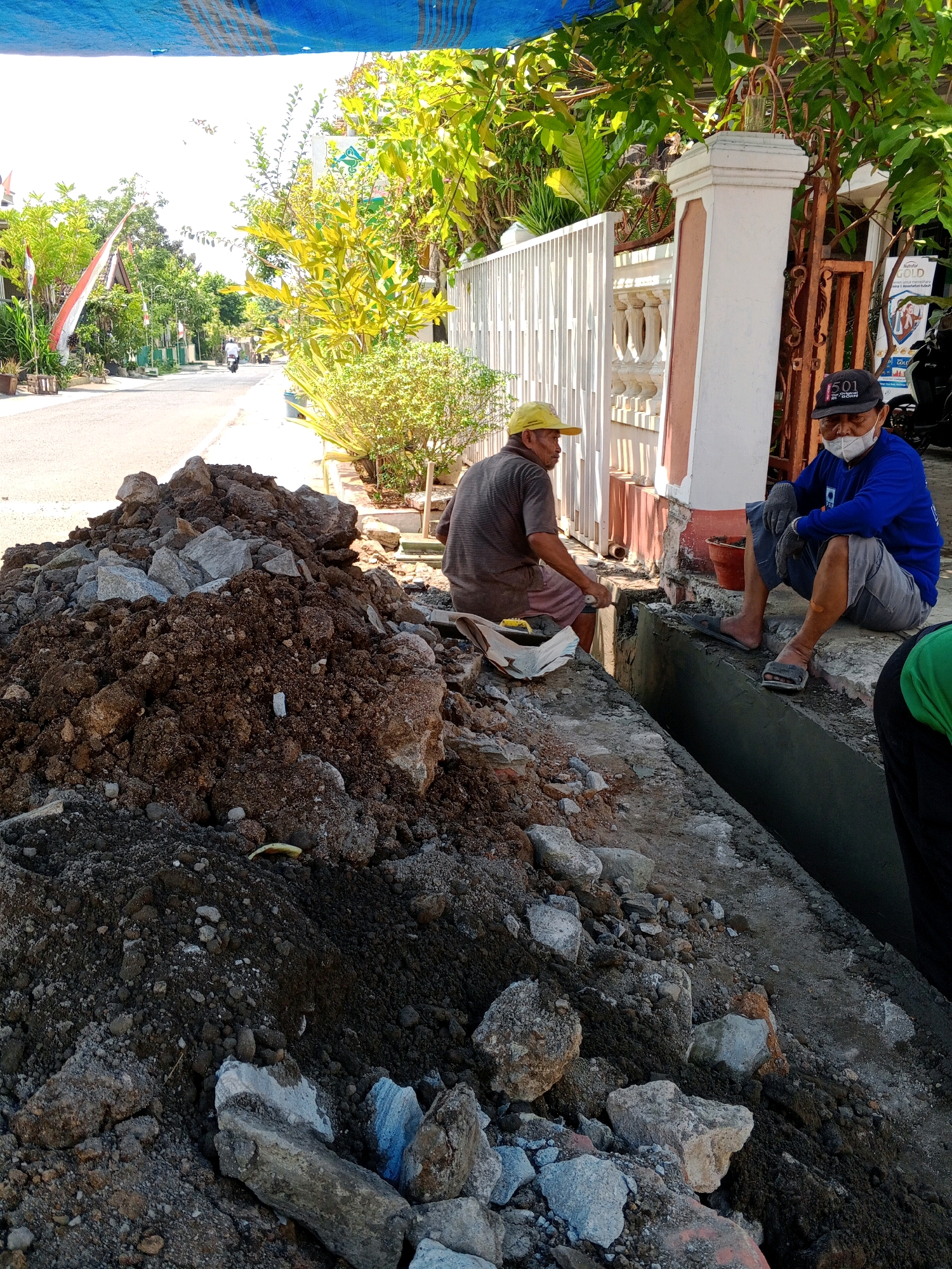In Giritirto, Indonesia, two men are engaged in construction work along a street. In the foreground, a large pile of dirt and rocks sits on the side of the road. Two men are working in a drainage ditch next to the pile. One man, wearing a yellow hat and a striped shirt, is sitting and appears to be mixing cement. The other man, wearing a blue long-sleeved shirt, shorts, a baseball cap, and a face mask, is sitting on the edge of the ditch. A portion of another person, wearing a green shirt, is visible in the lower right corner of the frame. The street is paved, and a white fence with decorative elements borders the property behind the workers. Trees and lush green foliage are visible on either side of the street.