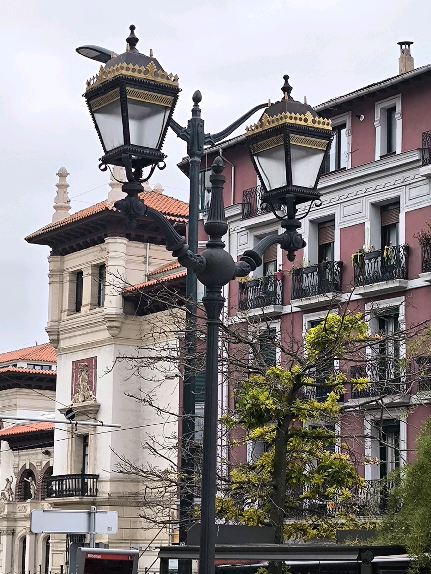 This image shows a decorative street lamp in front of buildings in Bilbao, Spain. The lamp is made of dark metal and features two ornate lampshades with gold trim and finials. Behind the lamp, a pink building with white trim and balconies is visible, as is a lighter-colored building with a tiled roof and arched windows. Bare tree branches, some with sparse green leaves, are in the foreground and middle ground. The sky is overcast. In the lower part of the image, a bus stop sign with "Bilbobus" text is present. The scene appears to be a street or plaza.