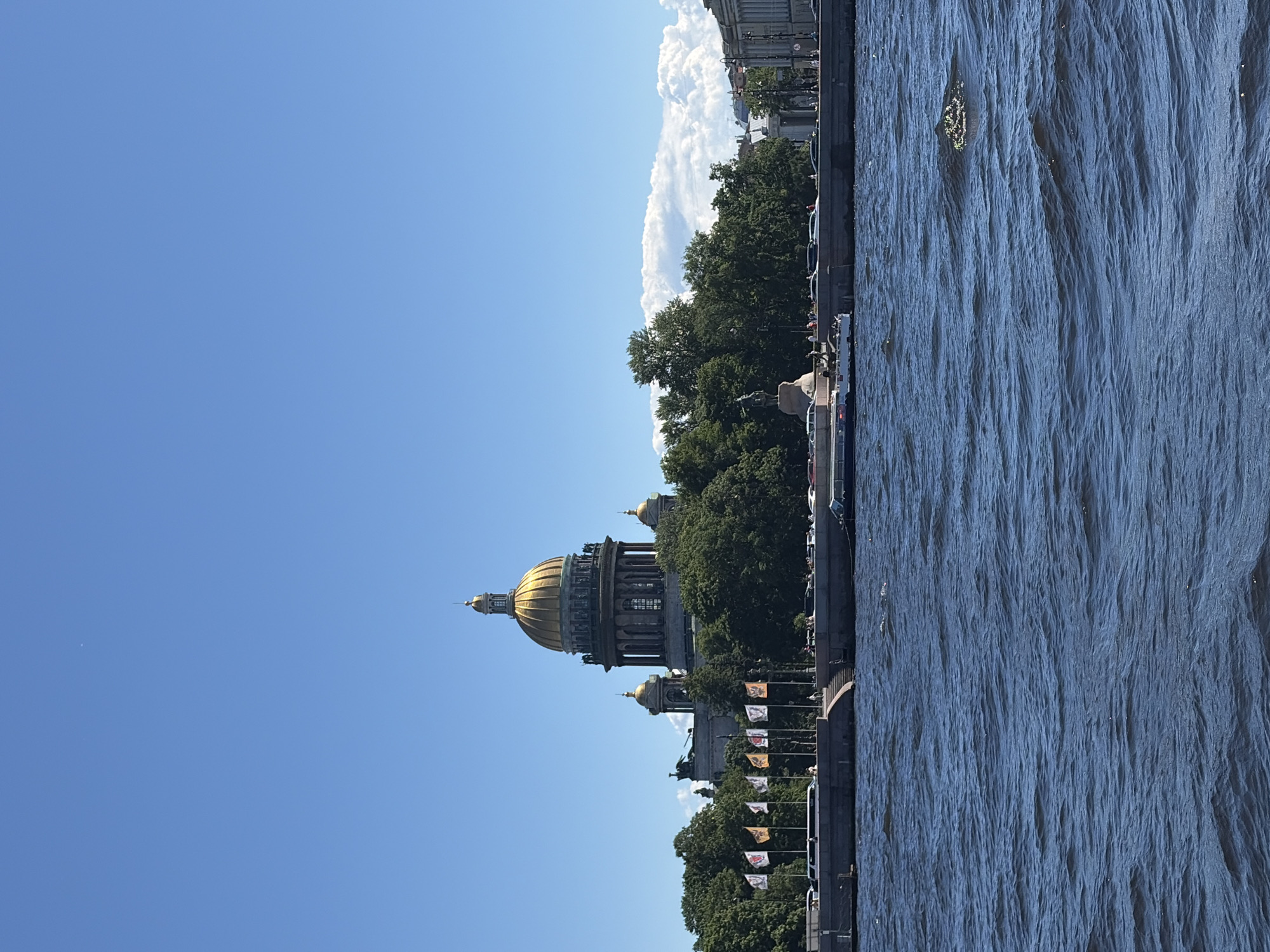 The image shows a body of water in the foreground, with a city skyline in the background. The sky is clear and blue. In the distance, a large building with a golden dome is visible, surrounded by trees. Flags are displayed on poles along the waterfront. Several boats are on the water, and there are people walking along the embankment. Cars are also visible on the street behind the embankment. The overall impression is of a sunny day in a city with a prominent architectural landmark. The building with the golden dome is St. Isaac's Cathedral in Saint Petersburg, Russia.