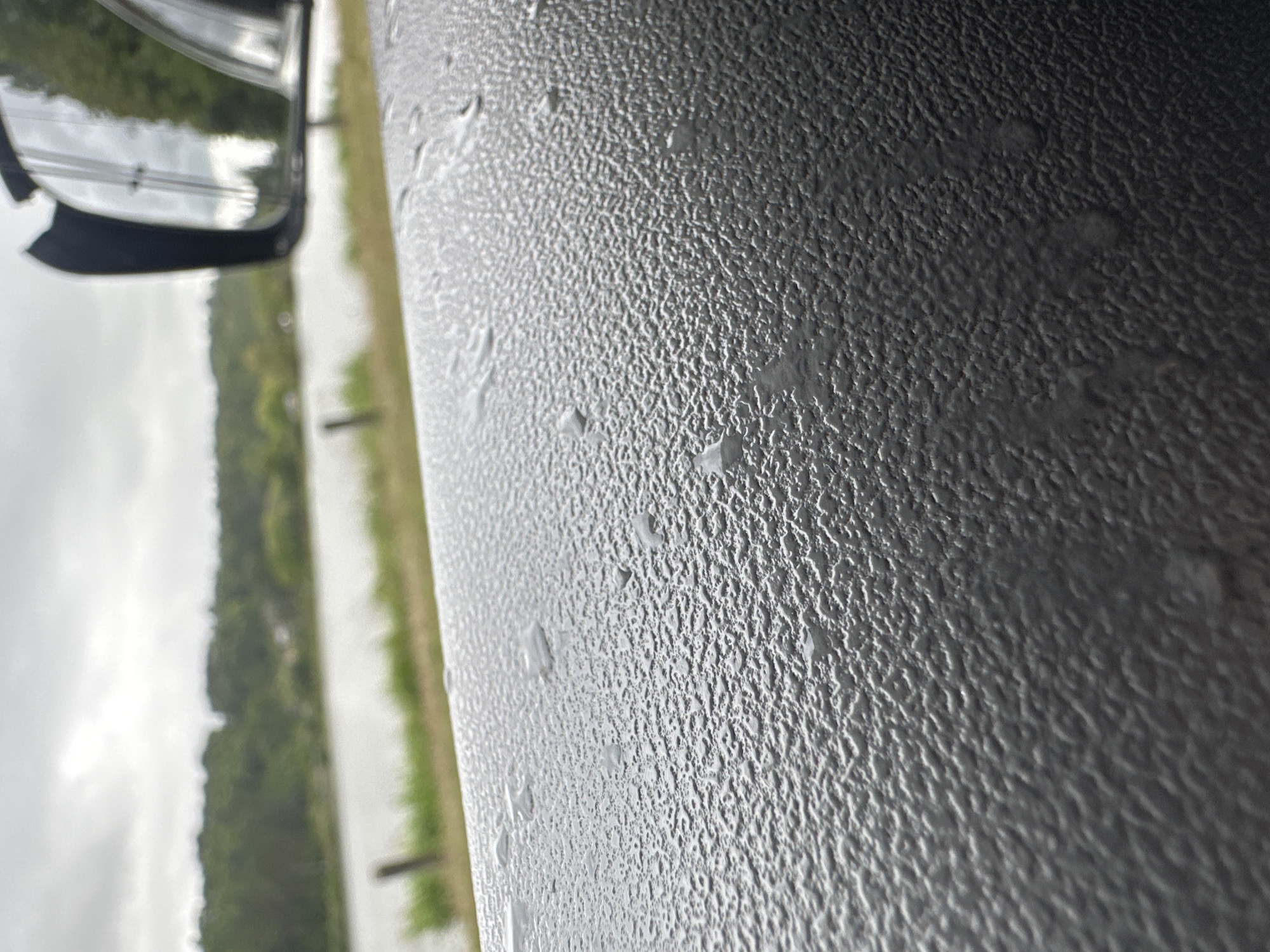 The image shows a close-up view of a textured, dark gray surface with water droplets on it, likely a car door or dashboard. In the background, out of focus, is a body of water, a grassy embankment, and a line of trees under an overcast sky. A side mirror of a vehicle is visible in the upper right corner, reflecting the sky and trees. The scene appears to be outdoors, possibly on the outskirts of a city given the natural elements in the background. No people or specific landmarks identifiable to Makiv, Ukraine are present in the image.