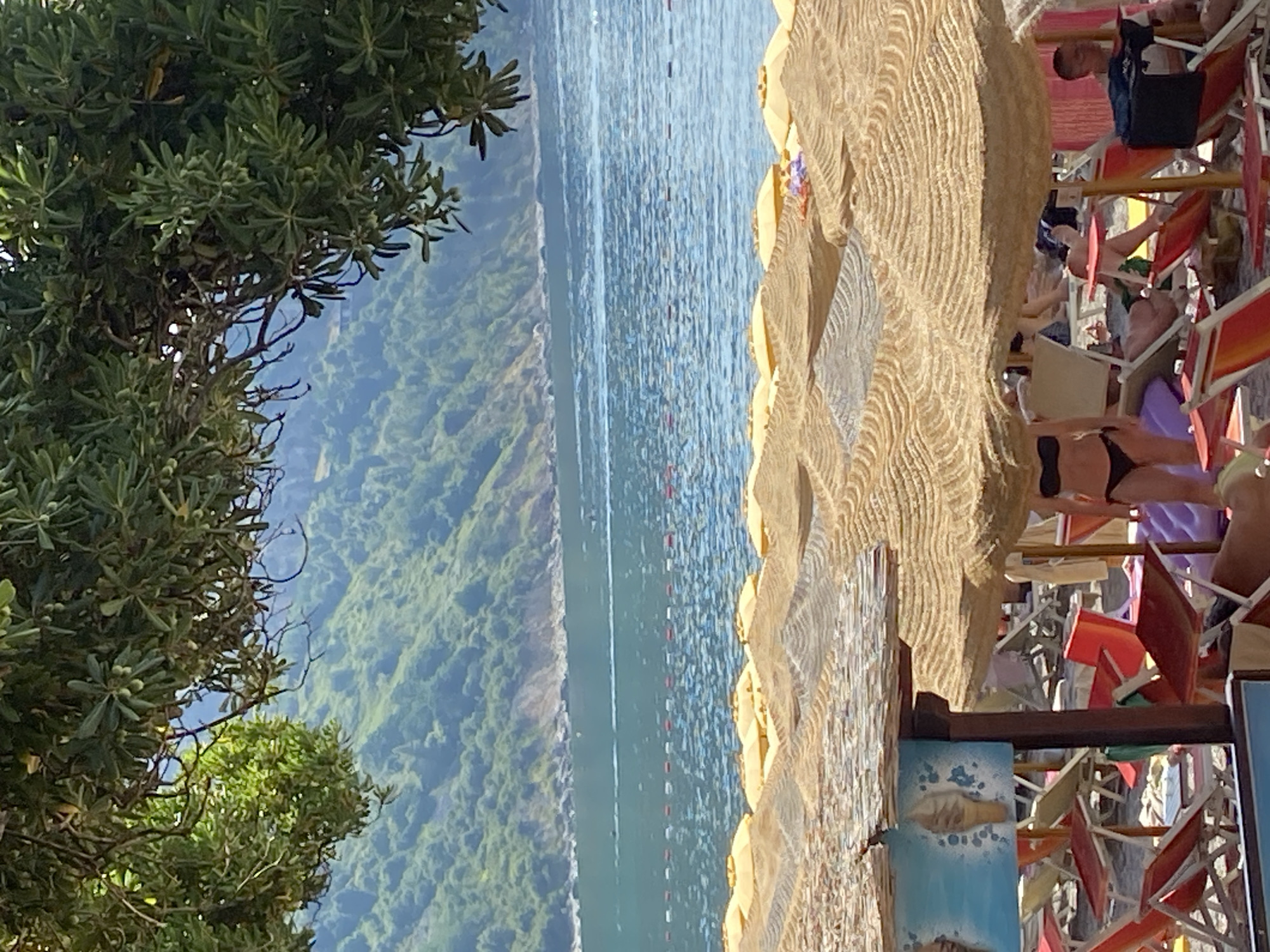 The image depicts a beach scene in Prijevor, Montenegro, on a sunny day. The foreground is dominated by straw umbrellas providing shade over numerous empty beach chairs, some of which are red and orange. A sign with an illustration of ice cream cones is visible on a wooden pole supporting one of the umbrellas. In the middle ground, the blue ocean stretches towards a lush, green, mountainous coastline in the background. A buoy line with red and white markers separates the swimming area from the open water. A woman in a black bikini is standing near the center, and other beachgoers are seated in chairs under umbrellas. The overall impression is one of a relaxed and scenic beach resort.
