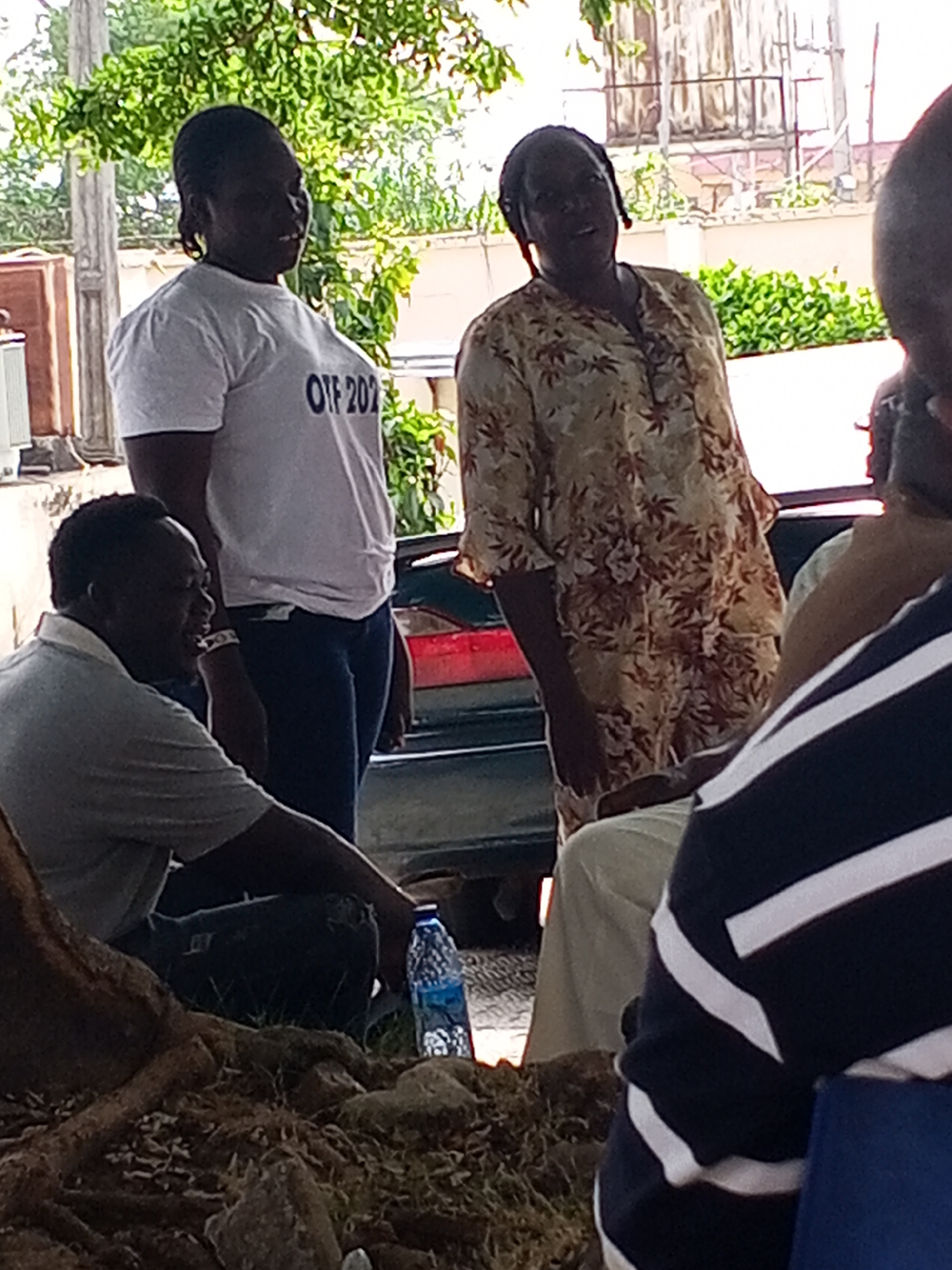 The image shows a group of people outdoors in Lokoja, Nigeria. A woman in a white t-shirt with "OTF 202" printed on it is standing next to another woman wearing a floral-patterned dress. A man wearing a gray shirt is sitting, and a bottle of water is on the ground near him. Another person is partially visible in the foreground wearing a striped shirt. Trees and plants are visible in the background.