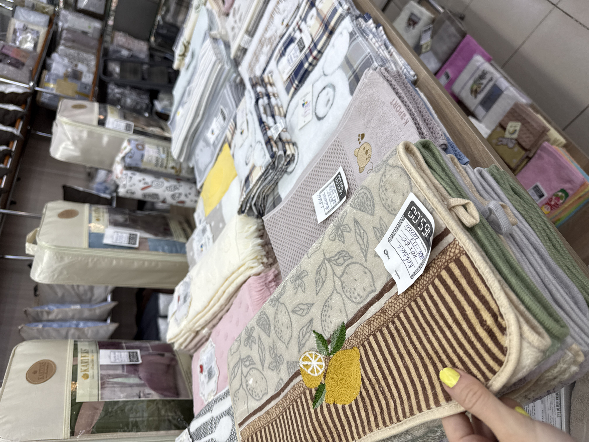 This image shows a stack of towels for sale in a store, presumably in the city of Chaikovsky, Russia, based on the text on the price tag in Russian and the word "Leurau" that can be read on the price tag. The towels are folded and stacked on top of each other on a shelf or display table. A hand with yellow nail polish is holding one of the towels, perhaps examining it before a potential purchase. Various other goods, possibly bedding sets or other home textiles, are visible on shelves behind the towels. The environment appears to be a retail store, possibly specializing in home goods or textiles.