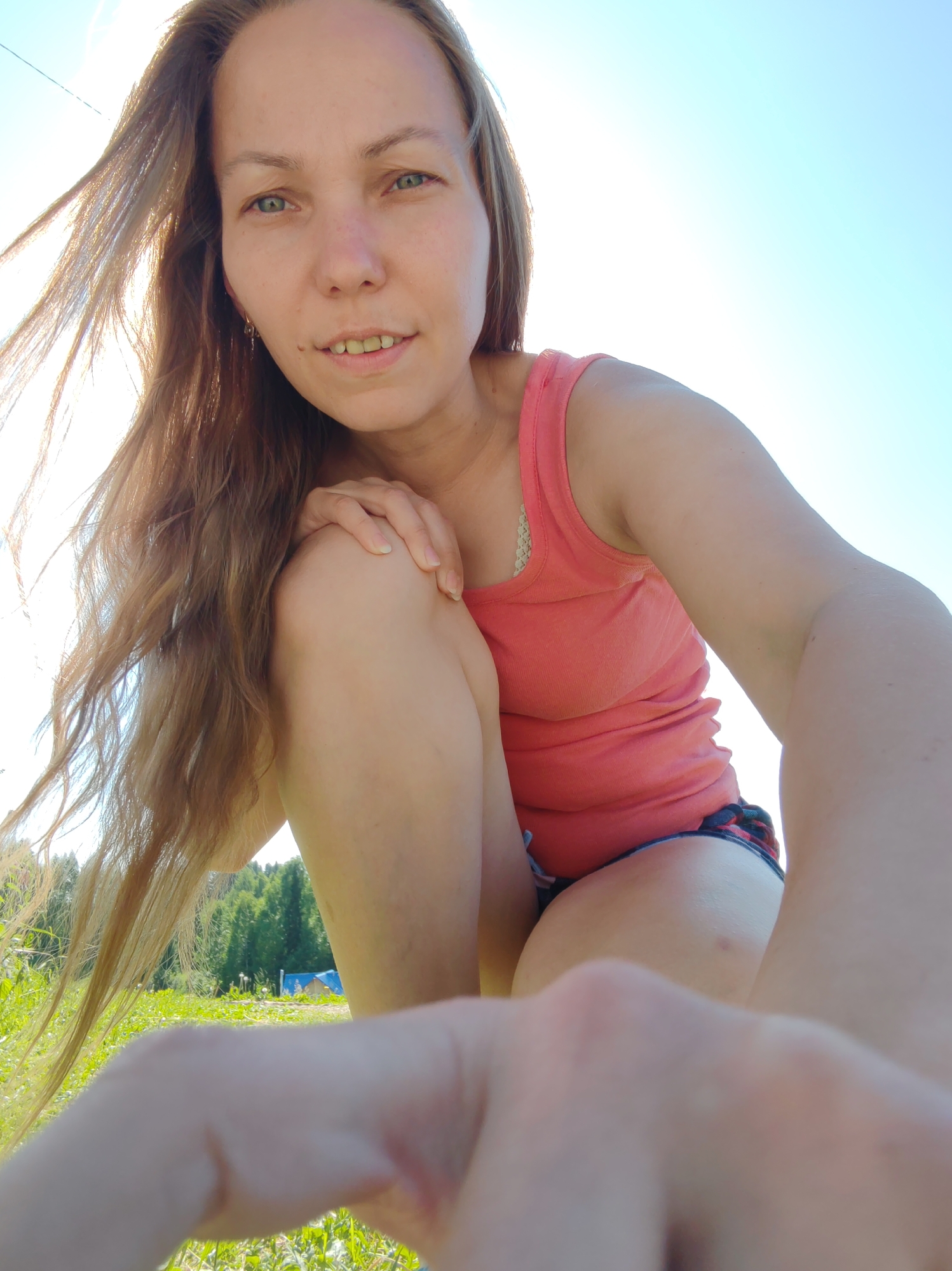 The image features a woman with long, wavy blonde hair, captured from a low-angle, first-person perspective. She is outdoors, seemingly in a grassy field with trees in the background under a clear blue sky, suggesting a daytime setting. The woman is wearing a coral-colored tank top and plaid shorts. Her right hand is placed on her left knee, and her left arm is bent, with her hand blurred in the foreground. Her expression appears relaxed and natural. The overall ambiance is casual and summery. The location is identified as Чухлэм, Russia.