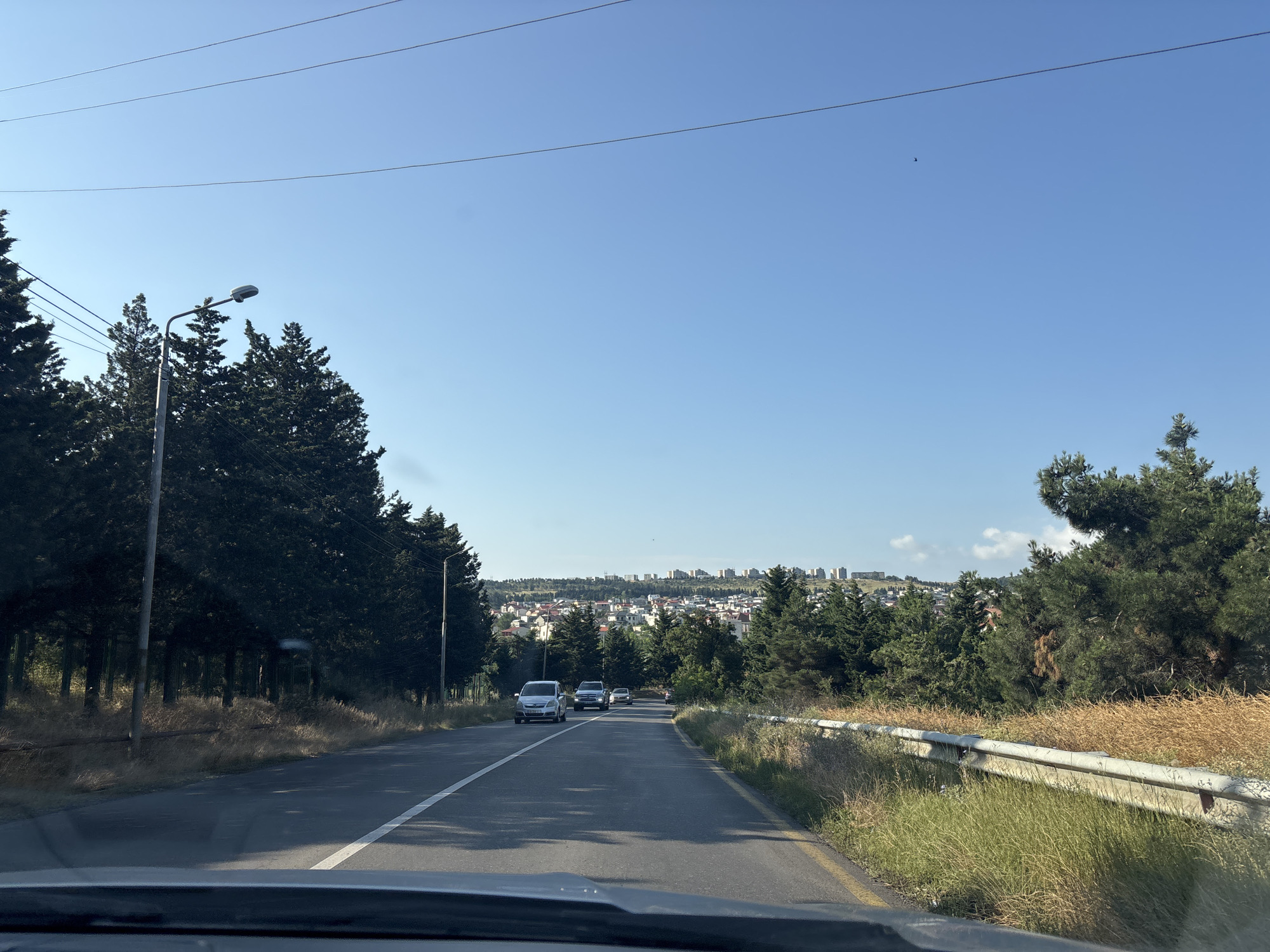 The image captures a view from a vehicle driving on a road in Tbilisi, Georgia. The road is paved with asphalt and has a white lane marker. On the left side of the road, there are tall, dark green coniferous trees and a street lamp. On the right side, there is a metal guardrail bordering dry, golden grass and some green vegetation. In the distance, a city skyline with buildings is visible, nestled among more trees. Several cars are driving on the road ahead, with one silver sedan and a white SUV being prominent. The sky is clear and blue, with thin power lines stretching across the upper part of the frame. The overall impression is of a suburban or semi-rural area leading into a city.