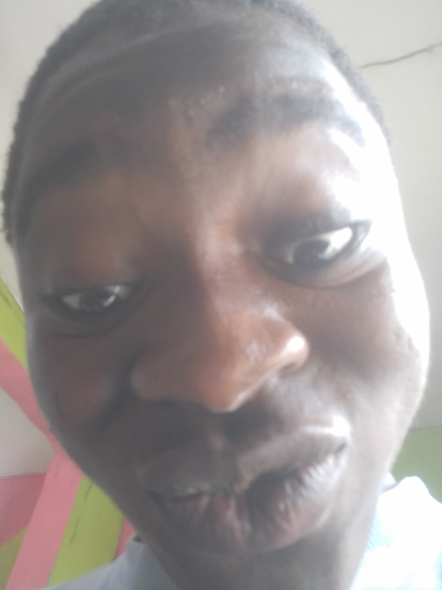 A close-up headshot of a dark-skinned person with short hair is shown. The person is looking slightly downward and towards the camera. The background is out of focus, but suggests an indoor setting with indistinct colors and objects. The exact location within the city of Unknown, Unknown is not discernible from the image.