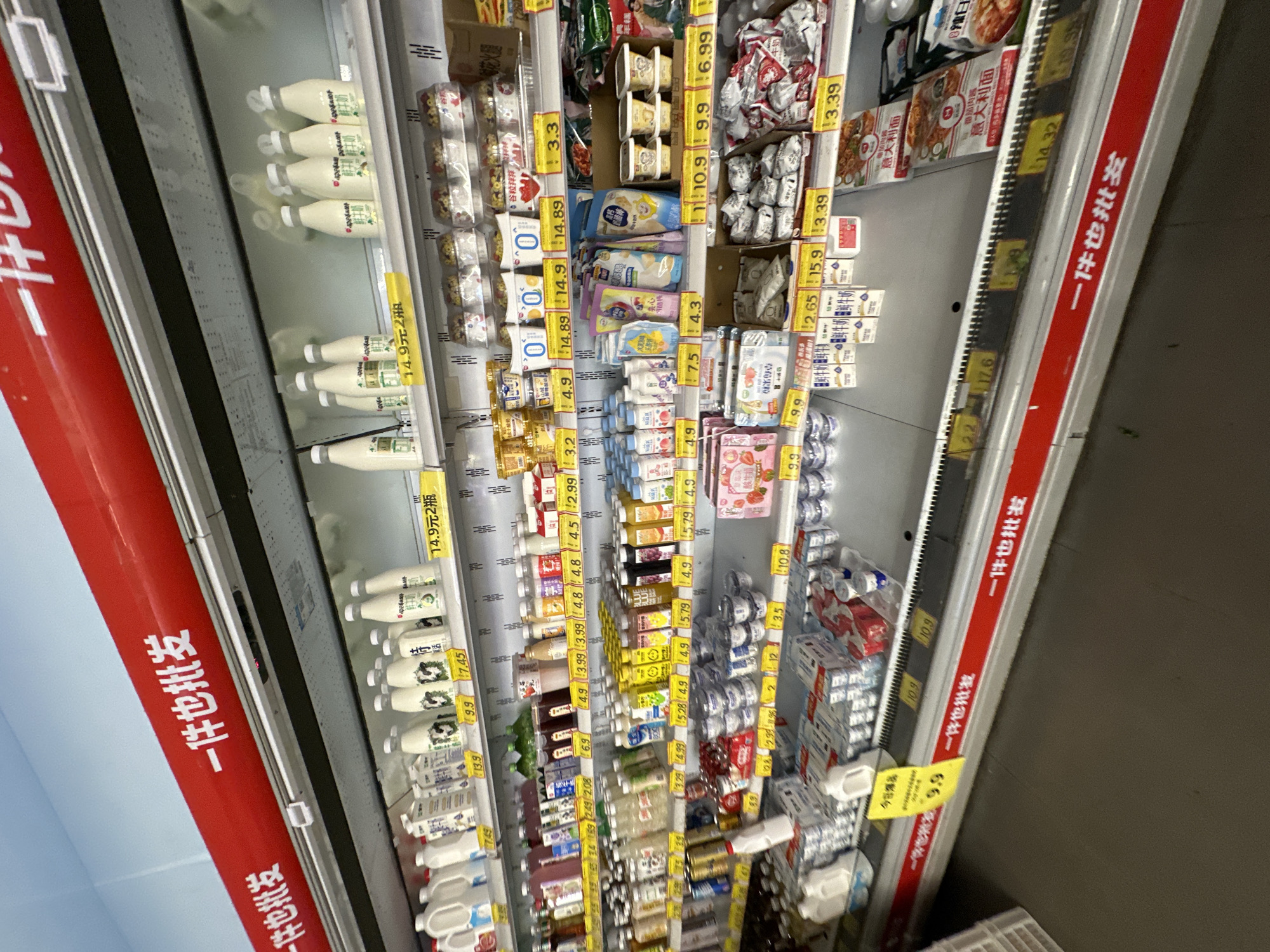 This image depicts a refrigerated display aisle within a supermarket or convenience store located in the Chongqing High-tech Zone, China. The scene is dominated by a multi-tiered, metal-shelved refrigeration unit fully stocked with various dairy products and chilled beverages.
Along the top and bottom edges of the display unit, prominent red banners with white Chinese characters read "一件也批发" (Yī jiàn yě pīfā), indicating that items can be purchased at wholesale prices even for single units.
The top shelf contains numerous white plastic bottles of milk or dairy beverages, some with green or black label accents. Visible yellow price tags indicate costs such as "14.9元2瓶" (14.9 RMB for 2 bottles), "13.9", "9.9", and "7.45" RMB.
The second shelf displays a denser arrangement of smaller bottles and cups, primarily yogurts, fermented milk drinks, and other chilled beverages. Several products are labeled with a "0", likely denoting a "zero" attribute (e.g., zero sugar or fat). Price tags on this shelf include "6.9", "4.9", "3.99", "4.8", "4.5", "2.99", "3.2", "14.89", and "3.3" RMB.
The third shelf features a diverse array of plastic bottles containing juices and flavored drinks in various colors (yellow, blue, pink), alongside packaged pouch drinks and small rectangular tubs of dairy or dessert items. Towards the left are strawberry-themed packaged goods. In the center and right, small individually wrapped items and bags, possibly containing cheese sticks or snacks, are visible. Prices here range from "3.29" to "15.9" RMB, including "4.99", "5.28", "4.9", "5.79", "7.5", "4.3", "10.9", "9.9", "6.99", "2.65", and "3.39" RMB.
The bottom shelf holds cartons of fresh milk (labeled "鲜牛奶"), additional small cups of yogurt or desserts, and pre-packaged frozen or chilled meals, specifically featuring "意式肉酱意大利面" (Italian meat sauce spaghetti). Price tags visible on this level include "10.9", "2.2", "17.6", and "14.32" RMB.
The environment is brightly illuminated by overhead fluorescent lighting. The floor is a plain, dark gray surface. No individuals are visible in the frame, and a white storage crate is partially visible on the far left. The products are neatly organized and aligned on the shelves.