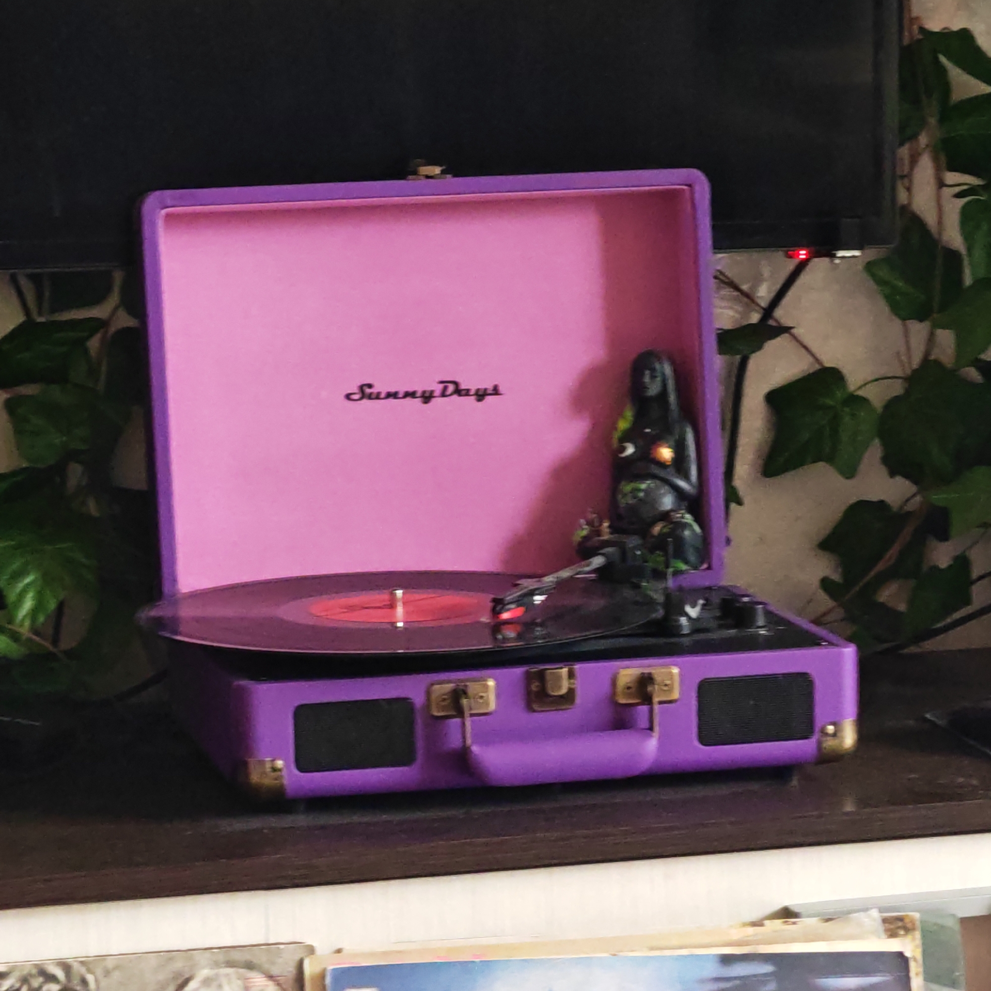 A vibrant purple portable record player, with its lid open, is the central focus of the image. The interior of the lid is a bright pink and features the script text "Sunny Days." A black vinyl record is placed on the turntable, with its red label visible. The tonearm is positioned over the record, suggesting music is either playing or about to be played.
To the right of the record player, a dark, statuette-like figure, possibly depicting a mermaid or goddess, sits upright. This figurine has light-colored or glowing accents on its chest and belly. The record player itself has brass-colored latches and a matching handle.
The record player rests on a dark wooden surface, possibly a shelf or table. Above it, a large black screen or television is partially visible, displaying a small red indicator light, suggesting it is powered on or in standby mode. Lush green leafy plants, possibly ivy, frame the left and right sides of the scene, trailing along a light-colored wall behind the electronic devices. The overall setting appears to be indoors. Below the wooden surface, parts of other items, possibly records or books, are visible, suggesting a decorated personal space. The scene does not indicate a specific time of day or particular weather conditions, and no human figures are present.