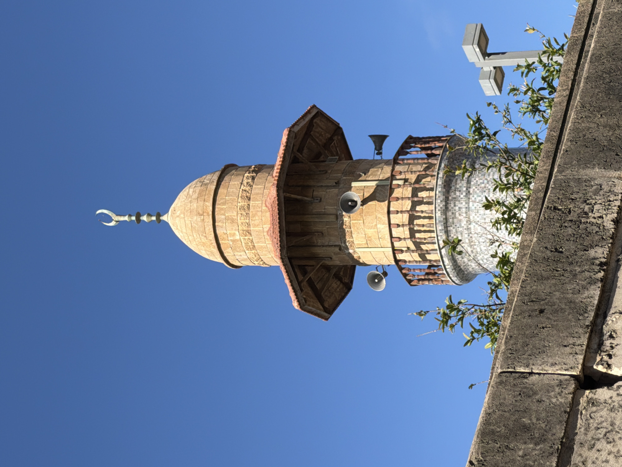 The image shows a minaret of a mosque against a clear blue sky. The minaret is made of light-colored stone and has a dome at the top, topped with a crescent moon. There are speakers mounted on the exterior of the minaret, which are used to broadcast the call to prayer. In the foreground, a concrete wall with some green foliage growing over it partially obscures the view of the minaret. To the right of the minaret, a modern street lamp is visible. This minaret is located at the Hassan Bey Mosque in Jaffa, a city that is now part of Tel Aviv-Yafo, Israel.