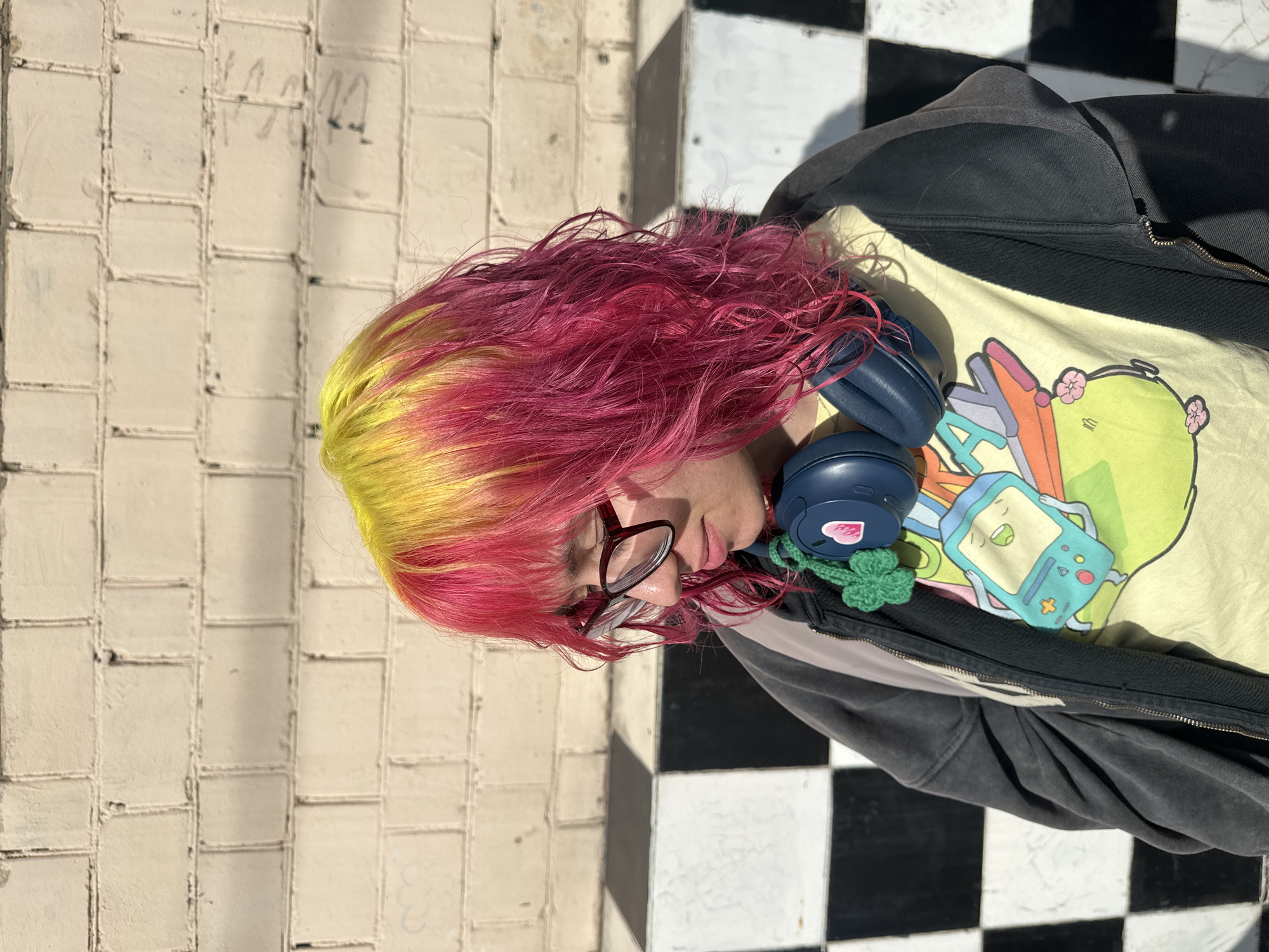 A young person with bright yellow and pink dyed hair, wearing glasses, a dark grey zip-up jacket, and a yellow t-shirt with a cartoon graphic, stands in front of a brick wall and a checkerboard pattern. They are wearing blue headphones around their neck, adorned with a green crocheted shamrock charm and a pink heart sticker. The person is looking downwards and to the side. The lighting suggests it is daytime, with shadows cast on the ground and the wall. The brick wall is light-colored and appears to be made of painted bricks. The checkerboard pattern is made up of black and white squares and forms a low structure or wall behind the person. The scene is set outdoors, possibly in an urban environment. The location is not identifiable as Moscow, Russia.