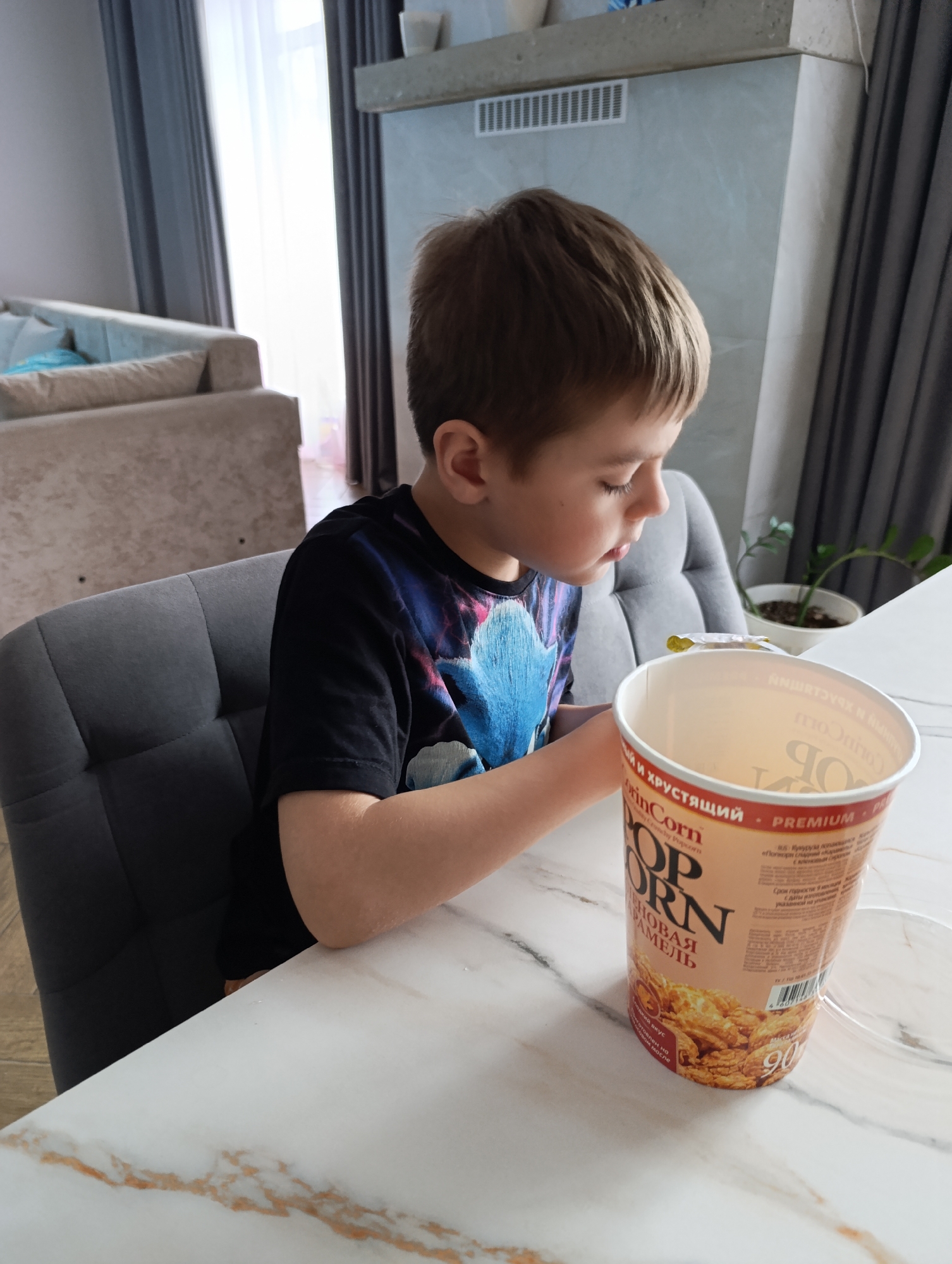 A young boy sits at a white marble table in what appears to be a domestic setting, possibly in an apartment. The child, facing slightly to the right, has short, light brown hair and is wearing a dark t-shirt with a colorful design. He is looking down at a tall, cylindrical container of "Pop Corn" which sits on the table in front of him. To the left, the boy is seated in a gray chair. The background includes a couch with blue pillows, a modern gray fireplace with decorative objects on top, and a window with gray curtains. There is also a potted plant visible. The location is identified as the city of Chaikovsky, Russia.
