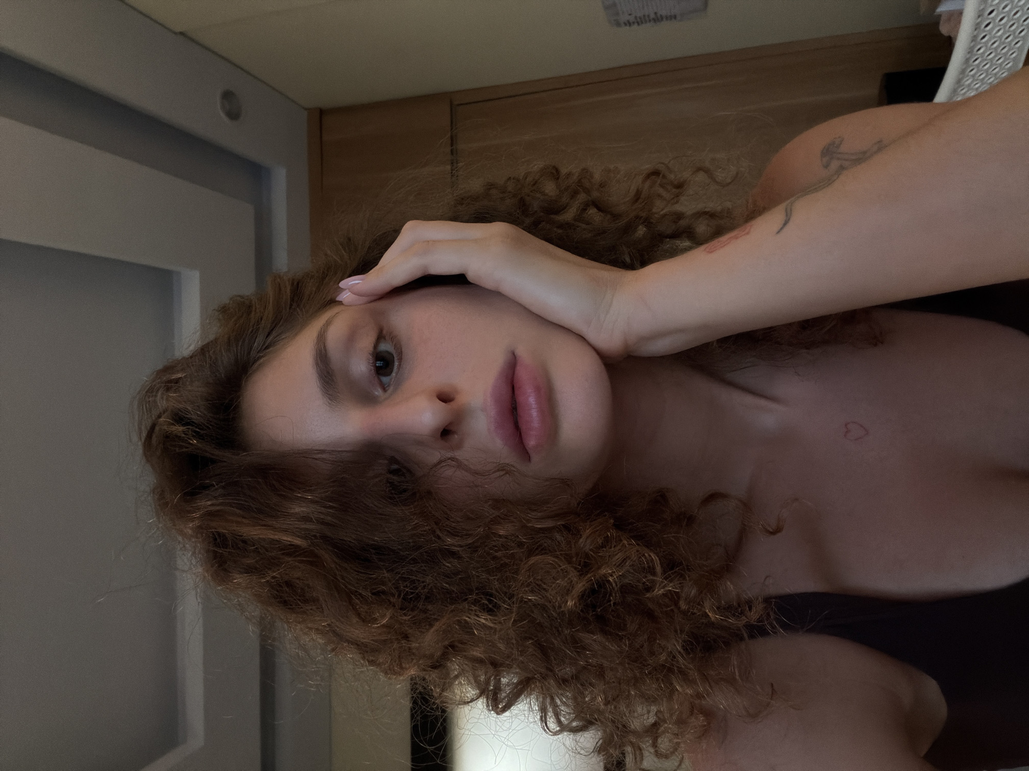 A medium-shot, rotated image displays a Caucasian woman with fair skin and light-brown, curly hair. Her left hand is resting on her forehead. The woman is looking directly at the camera. Tattoos are visible on her left arm and chest. The background includes a gray door, wooden panels, and a glimpse of a white plastic chair. The location is not discernible as North Kuta, Indonesia.