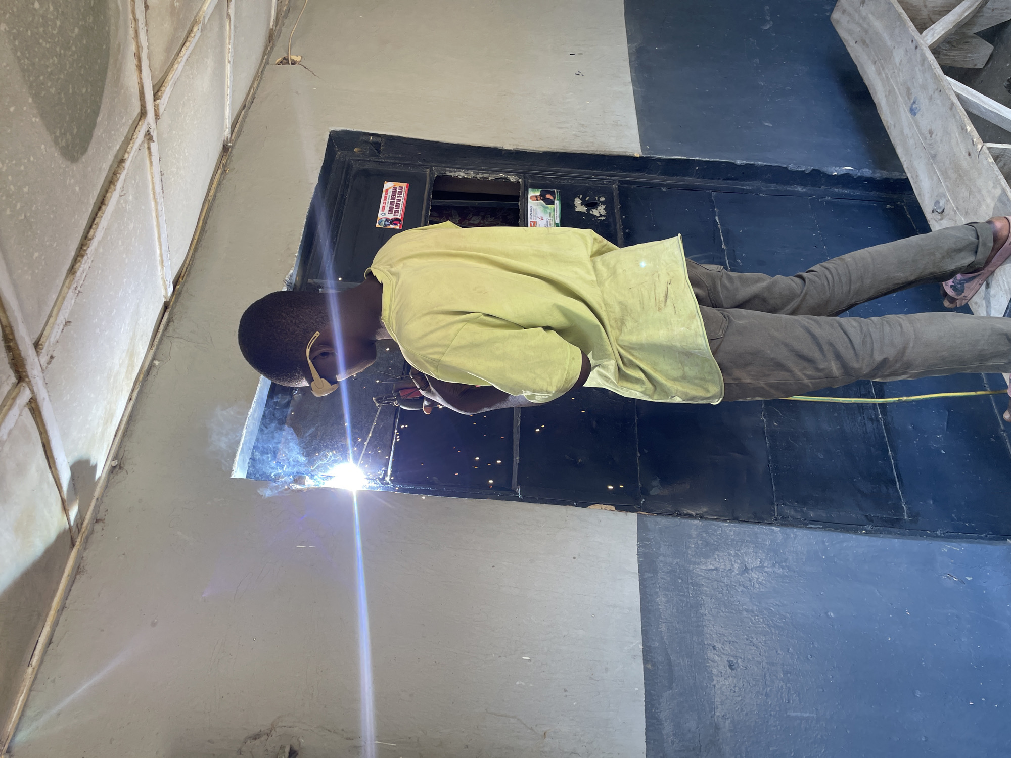 A person is welding metal within the frame of a window. The welder wears eye protection and a yellow shirt. The window appears to be set into a concrete wall. The location is Jos, Nigeria. A wooden ladder is visible in the upper right corner.