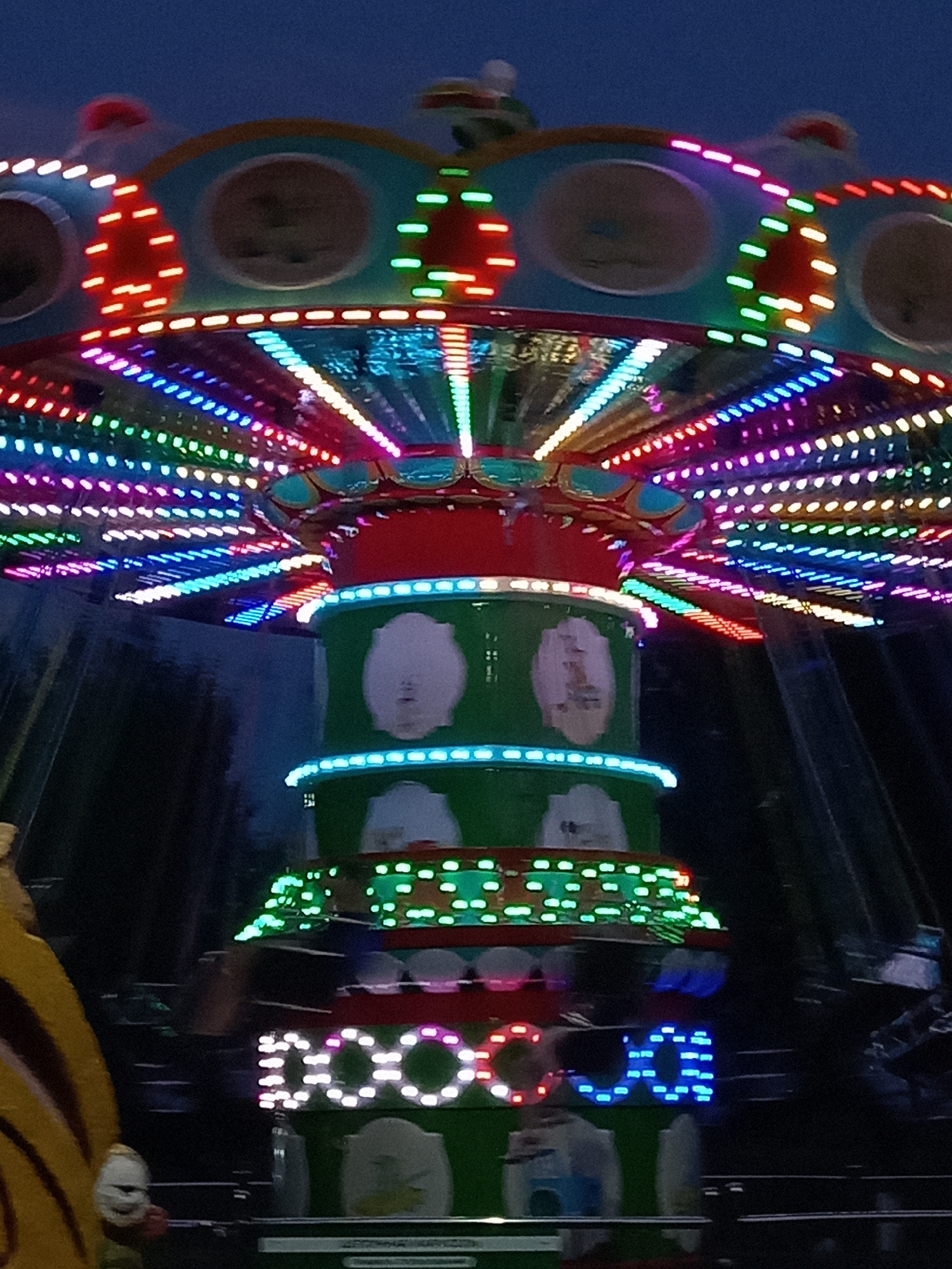 This is a close-up, low-angle shot of a brightly lit amusement park ride at dusk or night, likely located in the city of Rudny, Kazakhstan. The ride features multiple tiers with vibrant LED lights in red, blue, green, pink, and yellow, arranged in circular and radial patterns. The central column of the ride is green with decorative white shapes and has bands of blue and green lights. The top of the ride has a teal-colored canopy with embedded lights. The background is dark, with the blurred shapes of trees and possibly other structures visible. A portion of another ride, yellow with black stripes, is visible on the left side of the frame, with a small, white, clown-like figure attached. The overall impression is one of a lively, illuminated amusement park attraction.