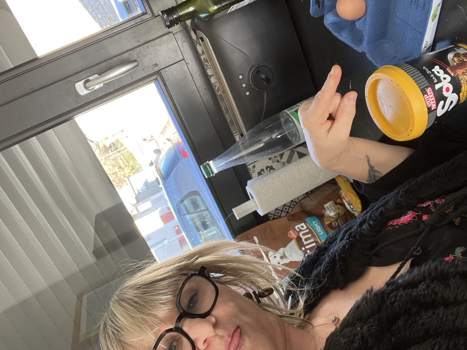 A female subject is depicted in a selfie-style photograph taken indoors, within a domestic setting in Chambœuf, France. The subject, with blonde hair and wearing black-framed glasses, is visible from the chest up, positioned towards the left of the frame. She wears a black textured outer garment over a dark top with pink and green embroidery, and a necklace with a small pendant. Her tongue is slightly extended, resting on her lower lip. Her right arm is raised and bent at the elbow, with her index finger extended and pointing downwards towards objects on a dark countertop. A dark tattoo, possibly of the Eiffel Tower, is visible on her right inner forearm.
On the dark countertop, from left to right, the following objects are present: a brown bag labeled "Purina Light" (partially visible), a white roll of paper towels on a stand, a clear plastic bottle with a green cap, a black toaster with a rotary dial and buttons, a dark green glass bottle with a black cap (likely containing olive oil), a blue-gray egg carton containing at least one brown egg, and a black "Nissin Cup Noodles Soba Wok Style Classic" container with a yellow lid.
Behind the counter, a window with a dark frame reveals an outdoor scene. Horizontal blinds are visible at the top of the window. Through the window, a dark-colored hatchback vehicle is parked on what appears to be a street or driveway, with residential buildings visible in the background under daylight conditions.