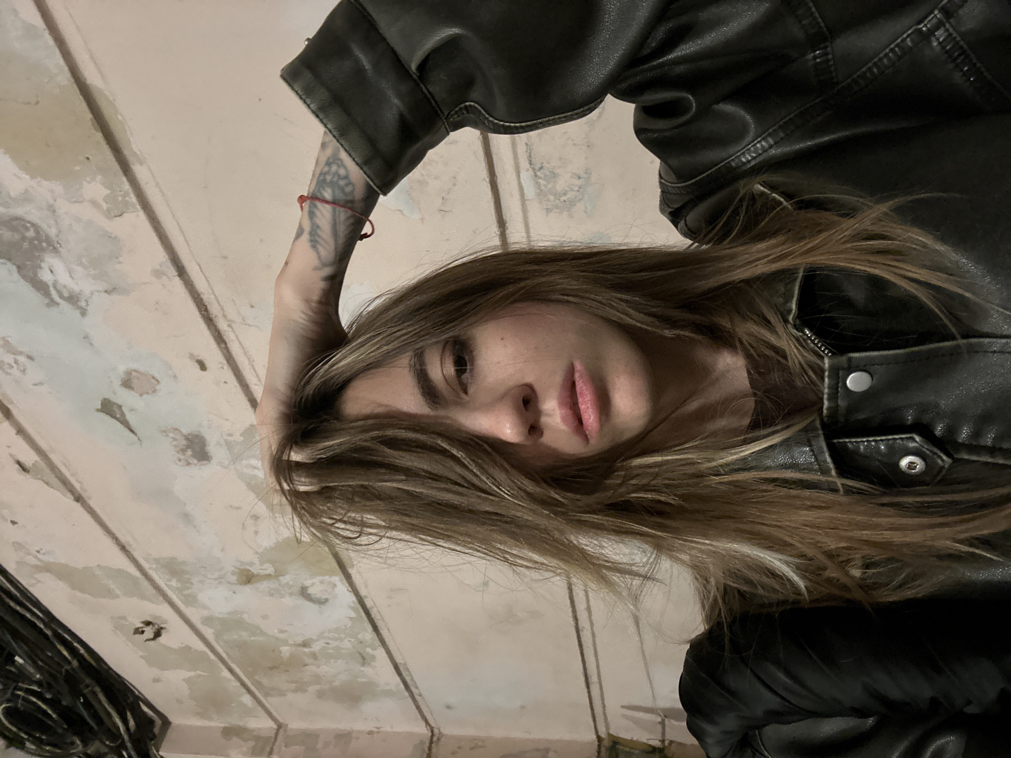 The image is a close-up selfie of a woman with long, light brown hair, wearing a black leather jacket. She is looking at the camera with her right hand tangled in her hair, and her left arm is bent with her hand resting on her head, revealing a tattoo on her forearm and a red string bracelet. The background appears to be a pinkish-beige wall with visible texture and some peeling paint, possibly in an indoor setting. Above the wall, a tangle of black wires is visible against a darker background. The lighting suggests an indoor environment, possibly at night or in a dimly lit space. The specific location in Moscow, Russia, cannot be determined from the provided image.