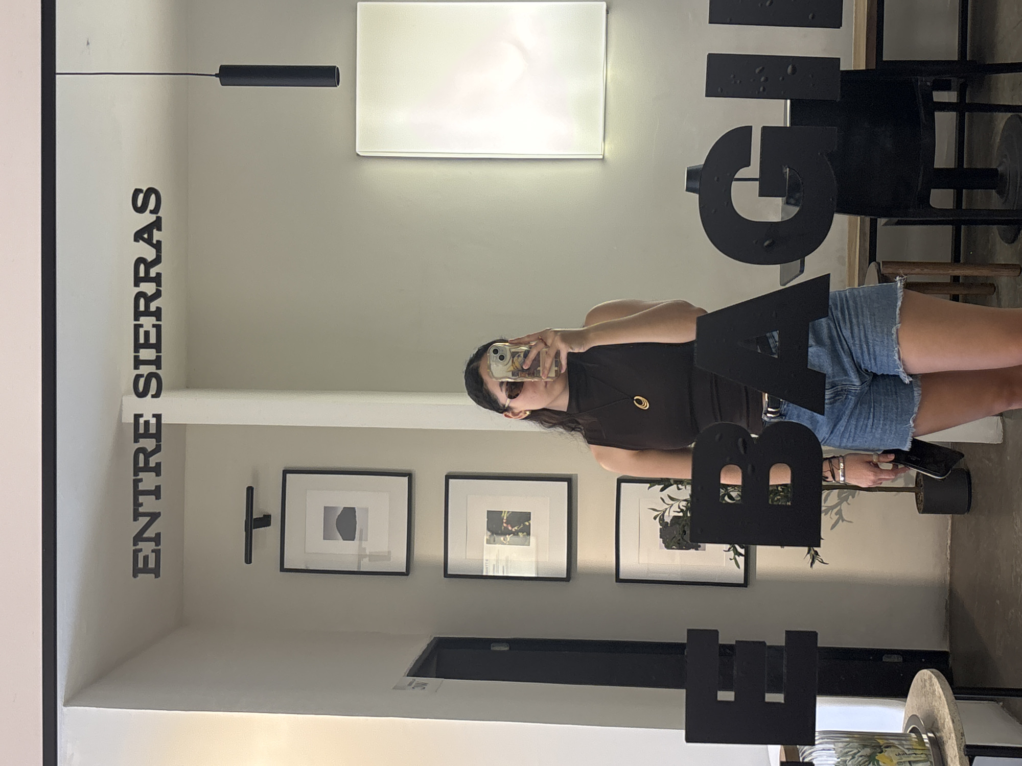 A vertically-oriented image depicts a mirror reflecting the interior of a commercial space in San Andrés Cholula, Mexico. The name "ENTRE SIERRAS" is vertically oriented, printed in black, on the wall to the left. The mirror reflects the rear of the space, where three framed pictures adorn the wall. The reflection also shows a woman with dark hair, sunglasses, a brown shirt, and denim shorts taking a selfie with a smartphone. The foreground of the reflection has three large, stylized black letters that appear to be a partial brand name or sign. Near the bottom of the reflection is a decorative plant. The space has neutral-toned walls and modern light fixtures.