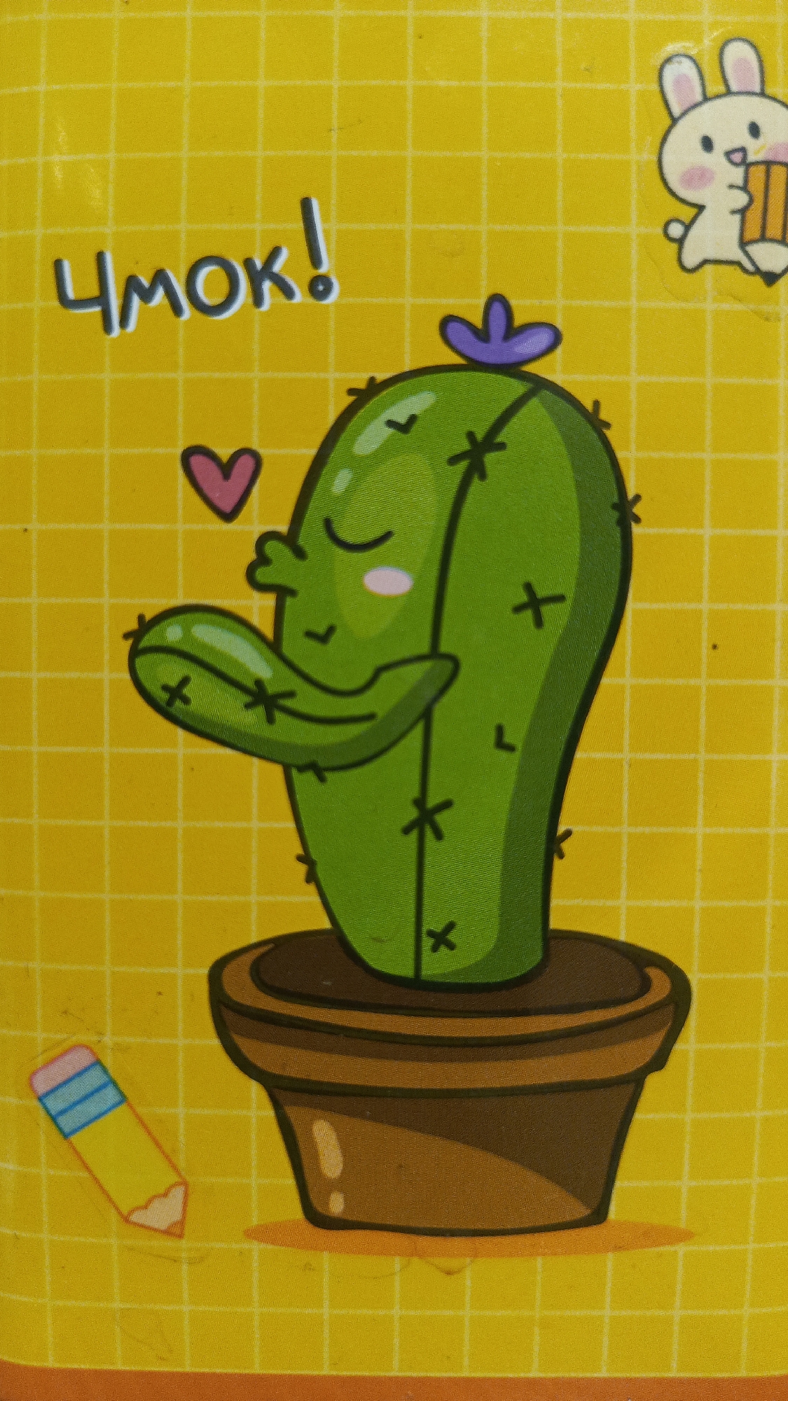 The image displays a cartoon of a cactus in a pot on a yellow, grid-patterned background. The cactus is green, with closed eyes and blushing cheeks, and has a purple flower on its head. It is also shown to be puckering its lips, as if to give a kiss, with a pink heart above its left arm. To the left of the cactus is a cartoon of a pencil with a pink eraser and blue and yellow segments. In the top right corner, there is a cartoon of a white rabbit holding a pencil. Above the cactus, the Cyrillic word "ЧМОК!" is written in large, dark letters, which translates to "Mwah!" or "Kiss!". There are no identifiable landmarks or elements in the image that would indicate a specific location in Zvenigorod, Russia.