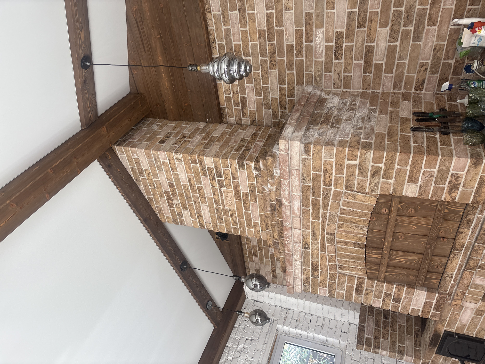 The image shows the interior of a room with a rustic design, featuring exposed wooden beams and a brick fireplace. The fireplace is made of light brown bricks with visible mortar lines and has a wooden door on its facade. There are three pendant lights hanging from the ceiling, each with a metallic shade. To the left, a wall is covered in white-painted bricks, and a window is visible. The ceiling is white and slopes upwards, supported by the wooden beams. The overall impression is of a cozy and decorative interior, possibly a living room or dining area. The location is identified as Georgiyevsk, Russia.