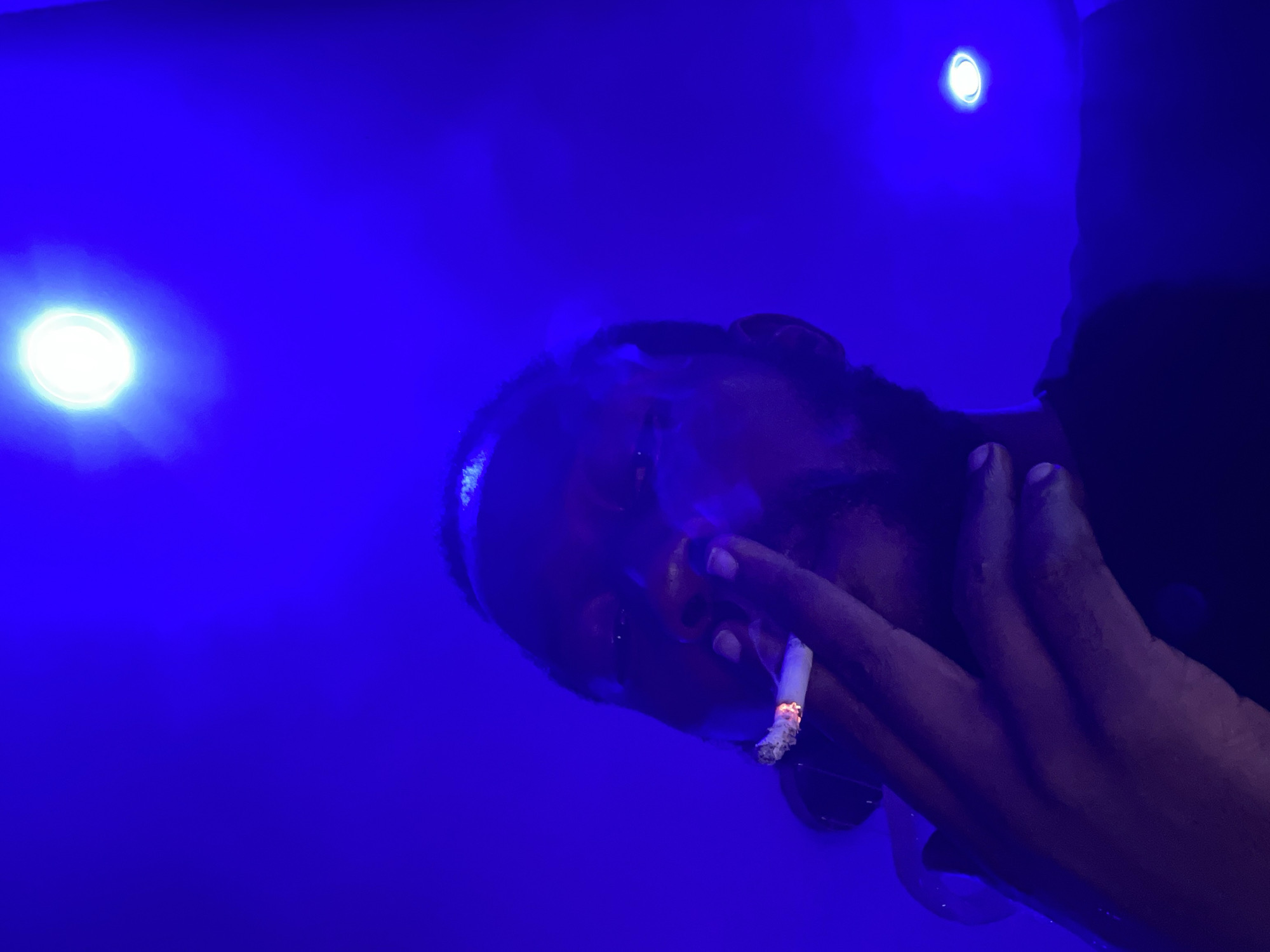 The image displays a person, likely of dark complexion, in a dark setting illuminated predominantly by a vibrant blue light. The person's head is tilted, and they are holding a lit cigarette between their fingers, visible through the blue hue. The glowing tip of the cigarette and a wisp of smoke are prominent. Two bright, circular light sources, emitting a white or pale blue glow, are visible in the background, contributing to the atmospheric lighting.
The intense blue light saturates the scene, casting a deep shade over the person's features and surroundings, making it challenging to discern specific details of their face, expression, or attire. However, a hand holding the cigarette is clearly visible near the face. The overall ambiance suggests a nighttime environment, possibly a club, bar, or lounge, typical of nightlife settings. The activity depicted is smoking, implying a moment of relaxation or contemplation. There is no visible text in the image. Given the context of Abuja, Nigeria, this scene likely portrays an indoor social or leisure activity.