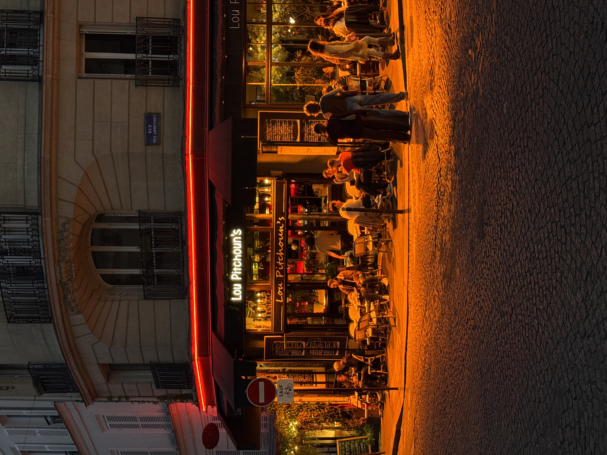 The image depicts a Parisian street scene at night. "Lou Pitchoun's", a restaurant with outdoor seating occupied by patrons, is situated on the right side of the frame. The street, constructed of cobblestones, is partially illuminated by the warm glow of the restaurant's lights. A building, featuring ornate window details and a street sign reading "Rue des Abbesses", stands to the left of the restaurant. A "no entry except for cyclists" sign is visible near the restaurant entrance. People are seated at the outdoor tables, engaging in conversation and dining.