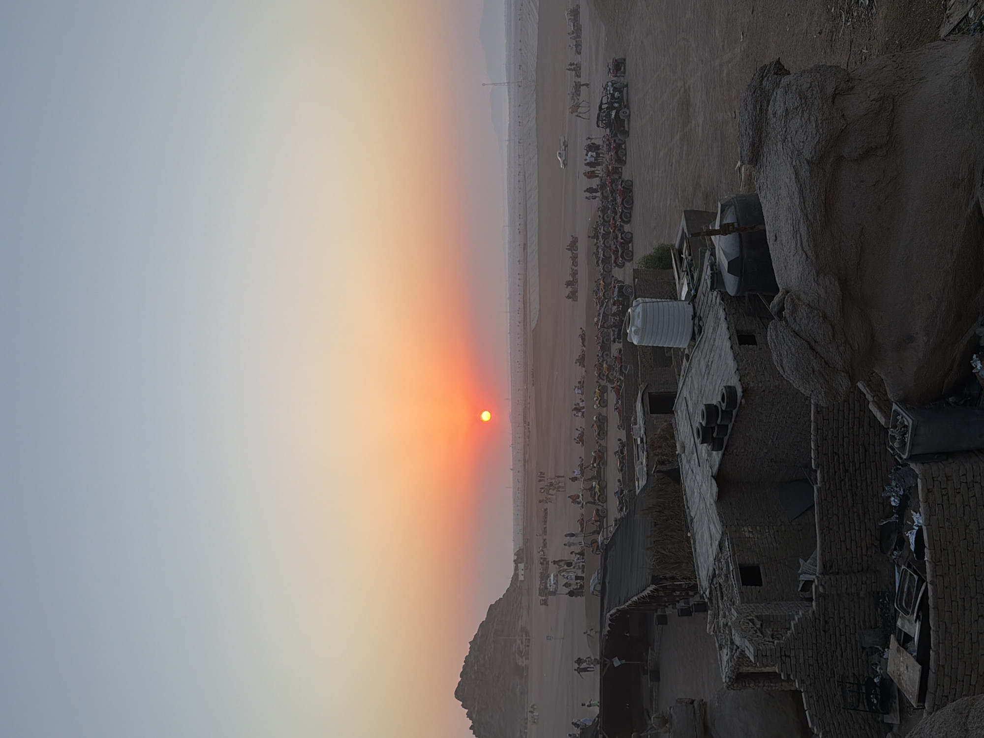 The image depicts a desert landscape at sunset, likely in Sharm El Sheikh, Egypt, given the context provided. The sky exhibits a gradient of colors, transitioning from a pale blue at the top to warm orange and red hues near the horizon, where the sun is setting as a small, bright disc.
In the mid-ground, a large gathering of people is visible, interspersed with numerous all-terrain vehicles (ATVs) and dune buggies parked in rows. These vehicles suggest an organized recreational activity, possibly a desert safari or tour. A white pickup truck and a camel with a rider are also present, further indicating a desert excursion. Several low-rise structures, appearing to be made of brick and possibly thatched roofing, are scattered across the terrain. One building in the foreground has a prominent white water tank on its roof and another darker tank next to it. The surrounding environment is sandy and arid, with a few rocky outcrops and a large, imposing rock formation on the left side of the image. The overall impression is of a popular tourist destination offering adventure activities in a desert setting during sunset.