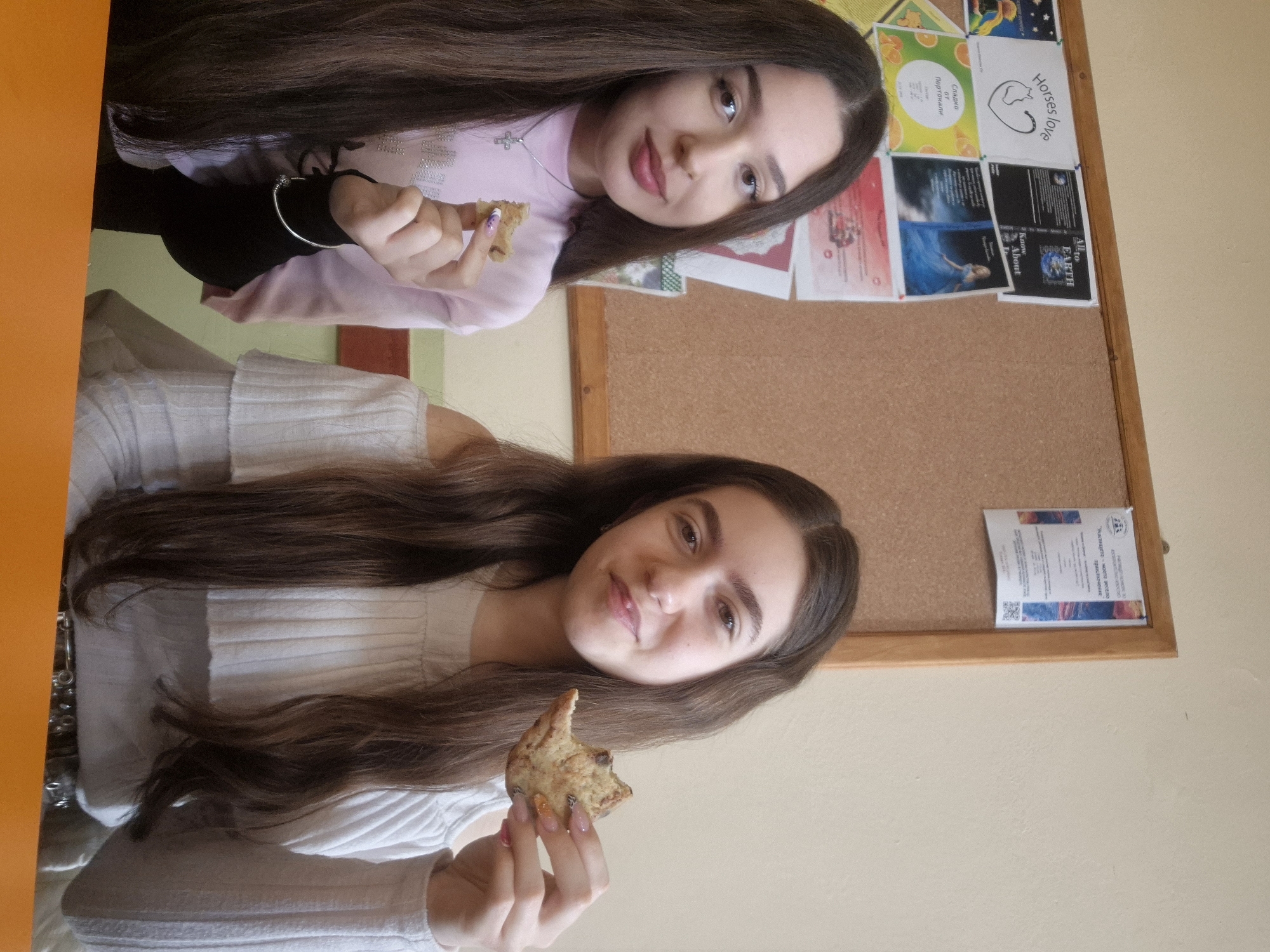 Two young women pose indoors, each holding a partially eaten cookie. The top woman is wearing a light pink long-sleeved shirt with a silver necklace and a bracelet on her left wrist. The bottom woman is wearing a white sweater. Both have long, dark hair. Behind them is a bulletin board displaying various papers and artwork. An orange object is visible on the left edge of the image. It is unknown if the location is in Pavlikeni, Bulgaria.