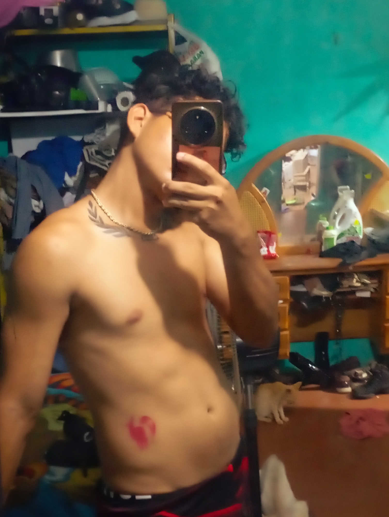 A shirtless person with curly hair and glasses is taking a selfie with a mobile phone, obscuring their face. The person has a gold chain with a leafy design, a leaf tattoo on the neck, and a red heart drawn on their lower abdomen. The background features a teal wall, a cluttered shelf with various items, and a vanity with a round mirror. A tan kitten is standing on the vanity. The photo was likely taken in a home in Altagracia, Venezuela.
