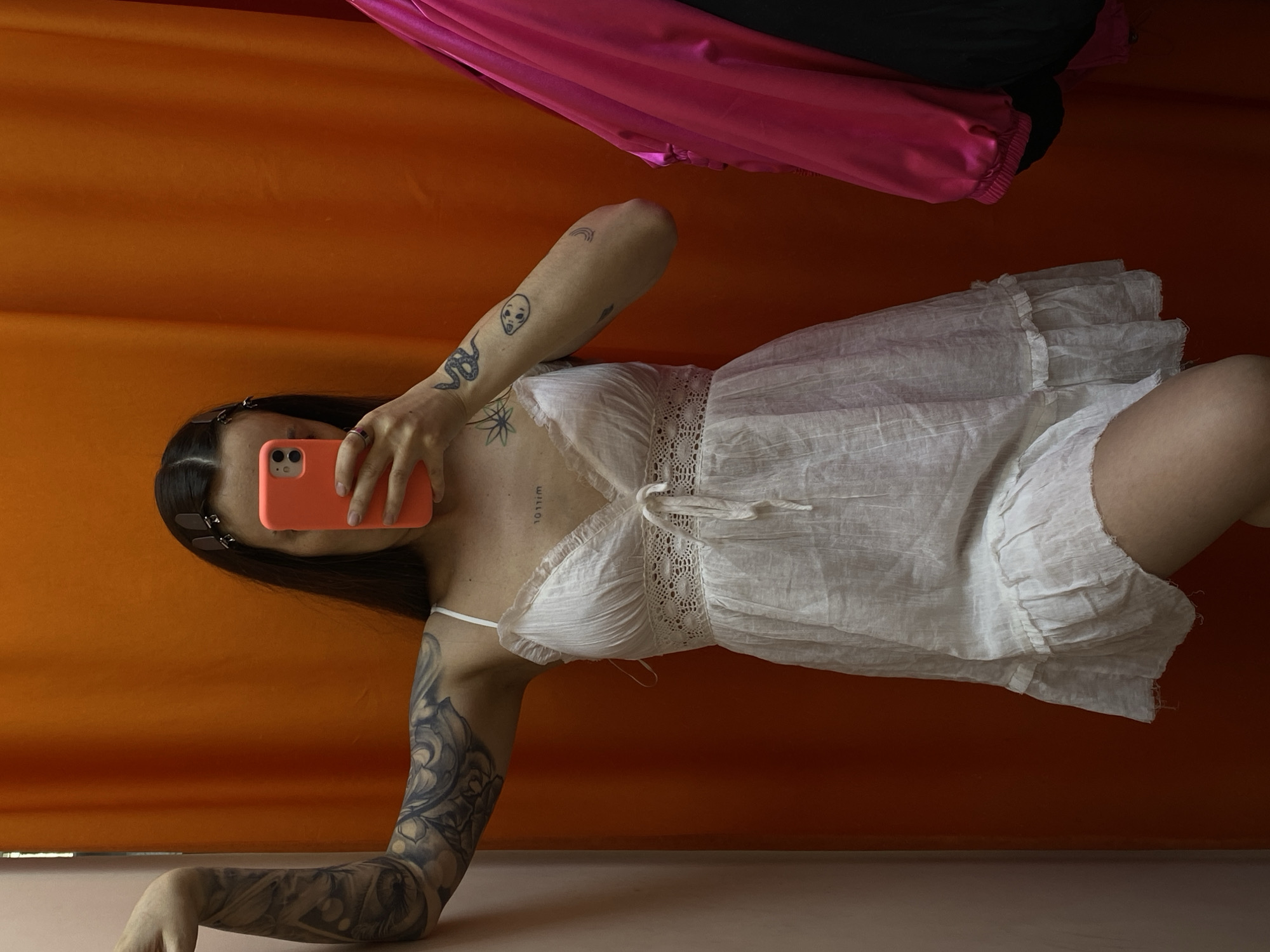 This image, set in Votkinsk, Russia, captures a person taking a self-portrait in a relaxed, reclined pose. The subject, likely female, has long dark hair held back with clips and is shown lying against a vibrant orange background, which appears to be a wall or curtain. She is wearing a light, white ruffled dress or nightgown featuring delicate lace detailing around the bodice and a tie-front closure. Her face is mostly obscured by a coral-colored smartphone she holds up, revealing only her chin and part of her mouth.
Numerous tattoos are visible on her arms and chest. On her right arm (viewer's left), a full sleeve tattoo with intricate, possibly floral or ornate, patterns covers the skin. Her left arm (viewer's right) displays several distinct, smaller tattoos, including a coiled snake, an alien head, and a rainbow. A small text tattoo reading "NOTHING" is visible on her upper chest, appearing upside down from the current image orientation. At the top of the frame, segments of shiny pink fabric and dark black fabric are visible, possibly draped clothing or decor. The bright lighting suggests a daytime indoor setting.