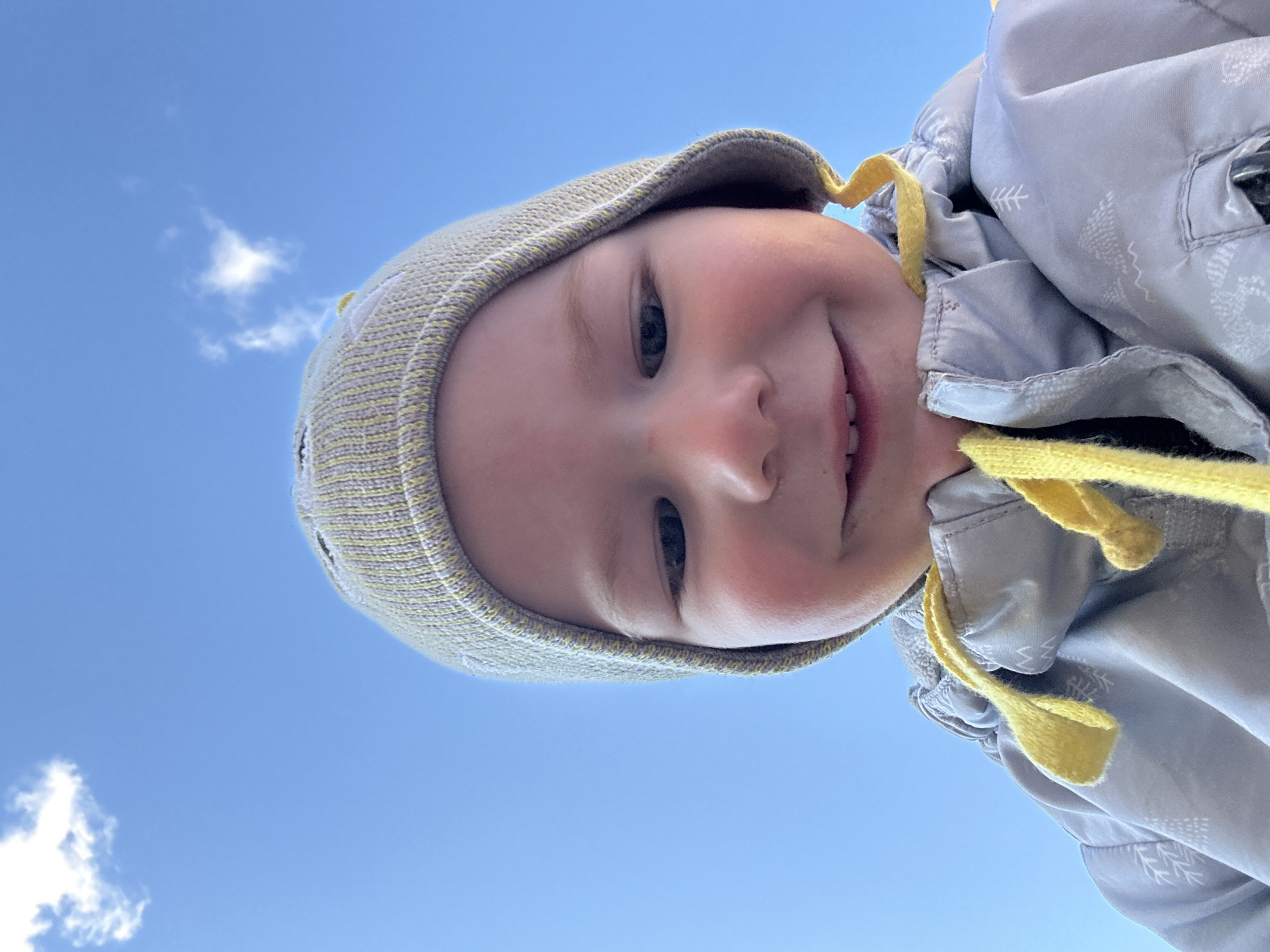 A low-angle shot depicts a young child wearing a gray knitted hat with yellow accents and a gray jacket, presumably standing outdoors. The child's face is turned towards the camera and a slight smile is visible. The background consists of a clear blue sky with a small cloud. The location is Прикамский, Russia.