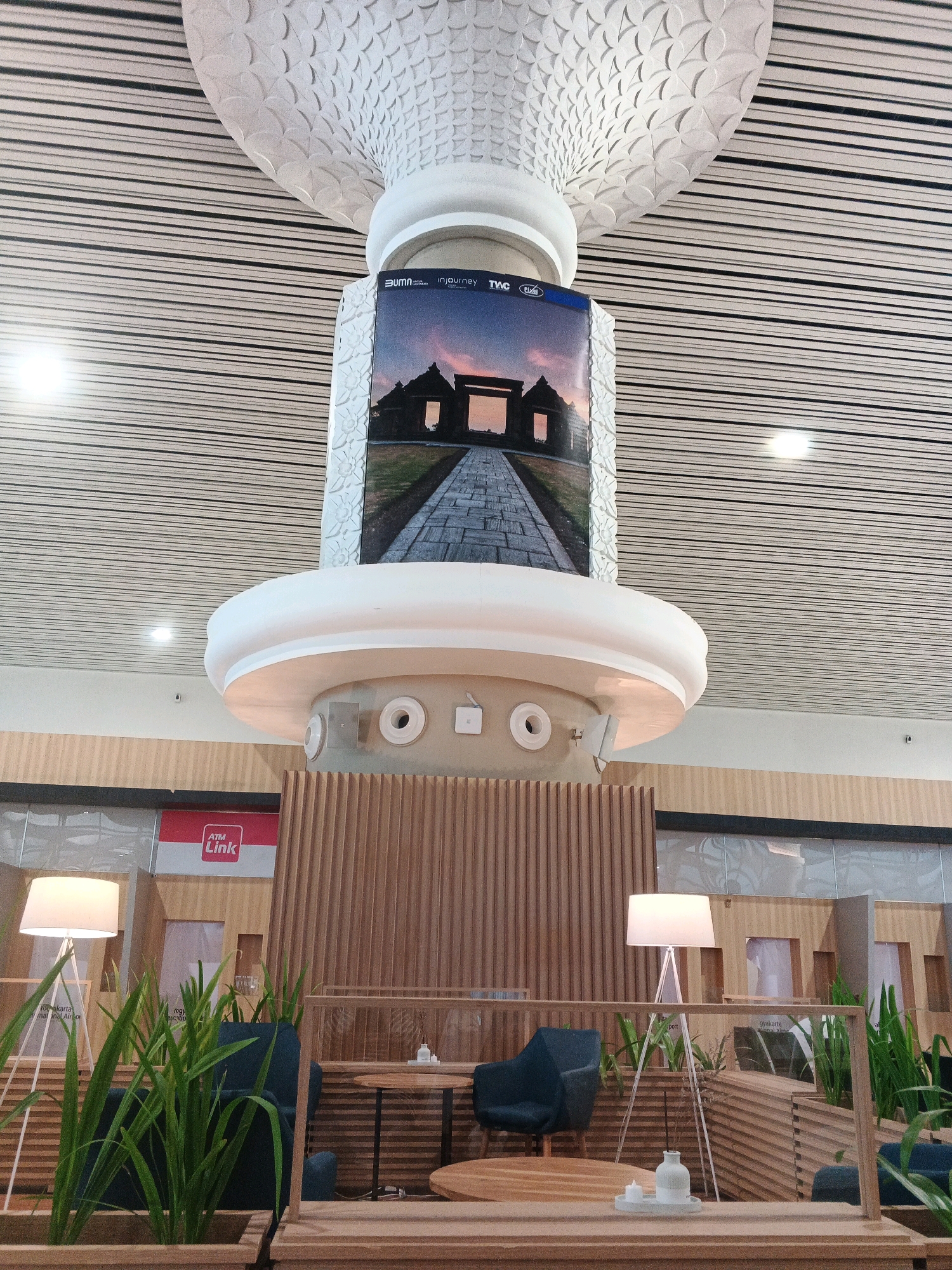 The image shows a modern indoor space, likely an airport or a large public building, with a distinctive ceiling made of horizontal wooden slats with black lines. In the center, a large, ornate white pillar rises, supporting a display screen showing an image of an ancient gateway with a stone path leading to it. The pillar has decorative carvings and a circular white element. Below this, a seating area is visible, featuring a dark blue armchair, a wooden table, and a floor lamp with a white shade. Green plants in wooden planters add a touch of nature to the scene. The background reveals more wooden paneling and glass partitions, with a sign for "ATM Link" in red and white. The scene appears to be in Kebonrejo, Indonesia, a location identified as part of the Indonesian context of the image. There are no people visible in the image.