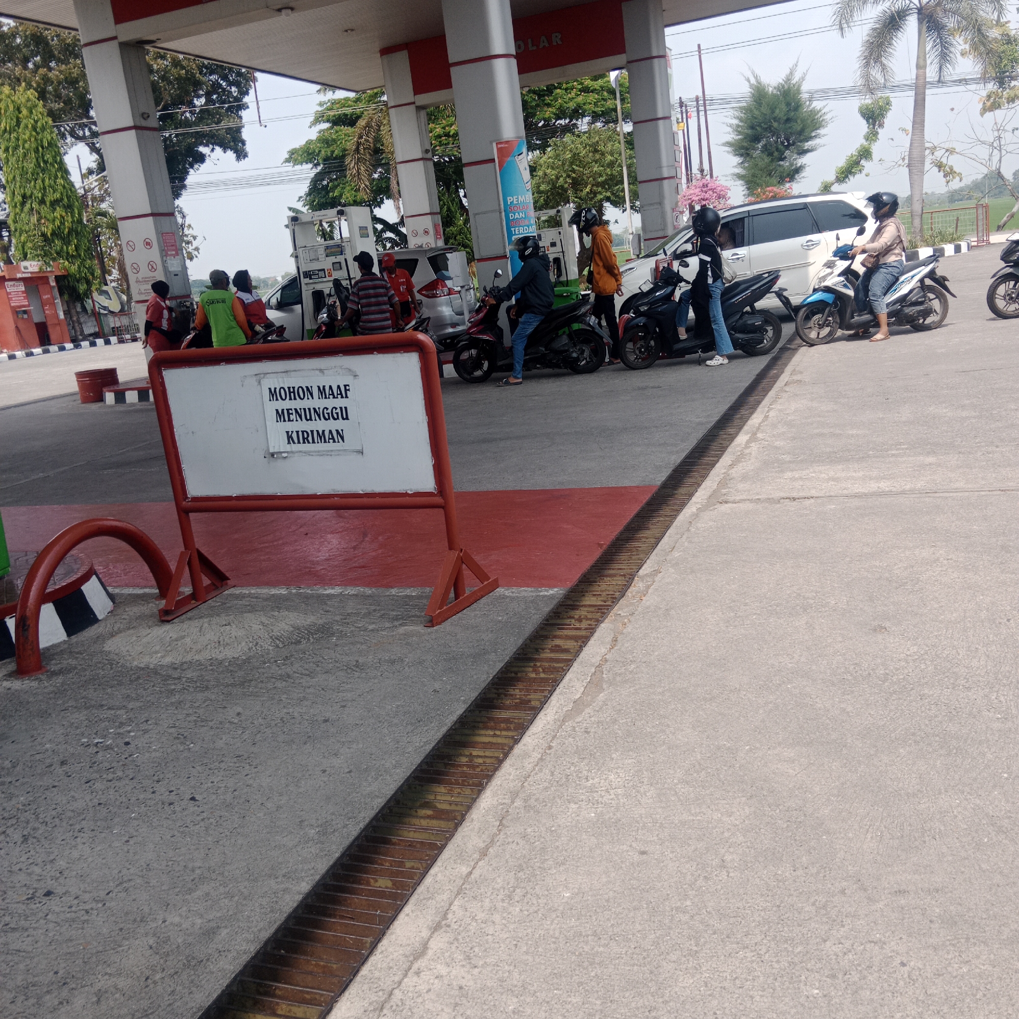 The image captures a scene at a gas station in Taraman, Indonesia. The foreground features a white sign with red trim, displaying the Indonesian text "MOHON MAAF MENUNGGU KIRIMAN," which translates to "SORRY, WAITING FOR DELIVERY." This sign is supported by a red metal frame. Adjacent to the sign, a red curved metal barrier is visible. The ground consists of concrete pavement with a metal grate drain running diagonally across it.
In the midground, several people on motorcycles are waiting. Some are employees of the gas station, identifiable by their uniforms (one in red and white, another in a green vest). Others appear to be customers. The gas station canopy is prominent, with red and white pillars supporting it. Gas pumps are visible, and some vehicles, including a white car and multiple motorcycles, are positioned around them. The background shows trees, a palm tree, and a glimpse of a cultivated field, suggesting a semi-rural or suburban setting. The sky is overcast. The overall atmosphere is one of waiting and service at a busy refueling point.