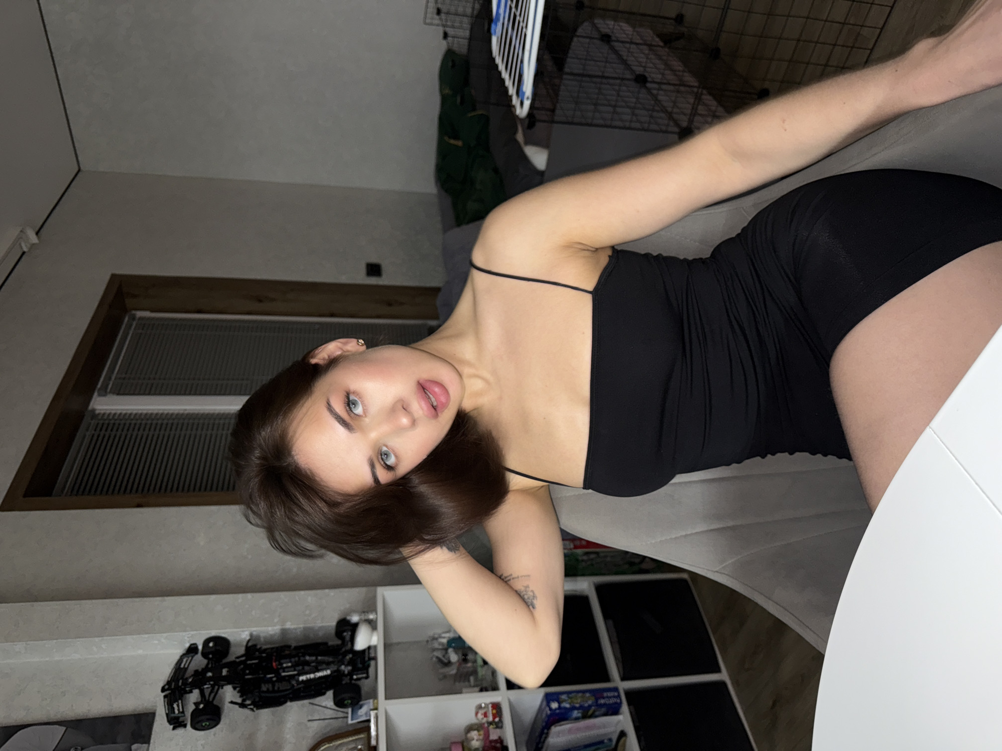 A young woman with fair skin and brown hair, wearing a black spaghetti strap dress, is posing for a picture indoors. She is lying on a gray upholstered surface, possibly a sofa or chair. Her right arm is raised, and her gaze is directed towards the camera. The room has a neutral color palette with a window in the background. On a shelf unit behind her, there is a toy car and various other items. The setting appears to be a residential space in Лобаново, Russia.