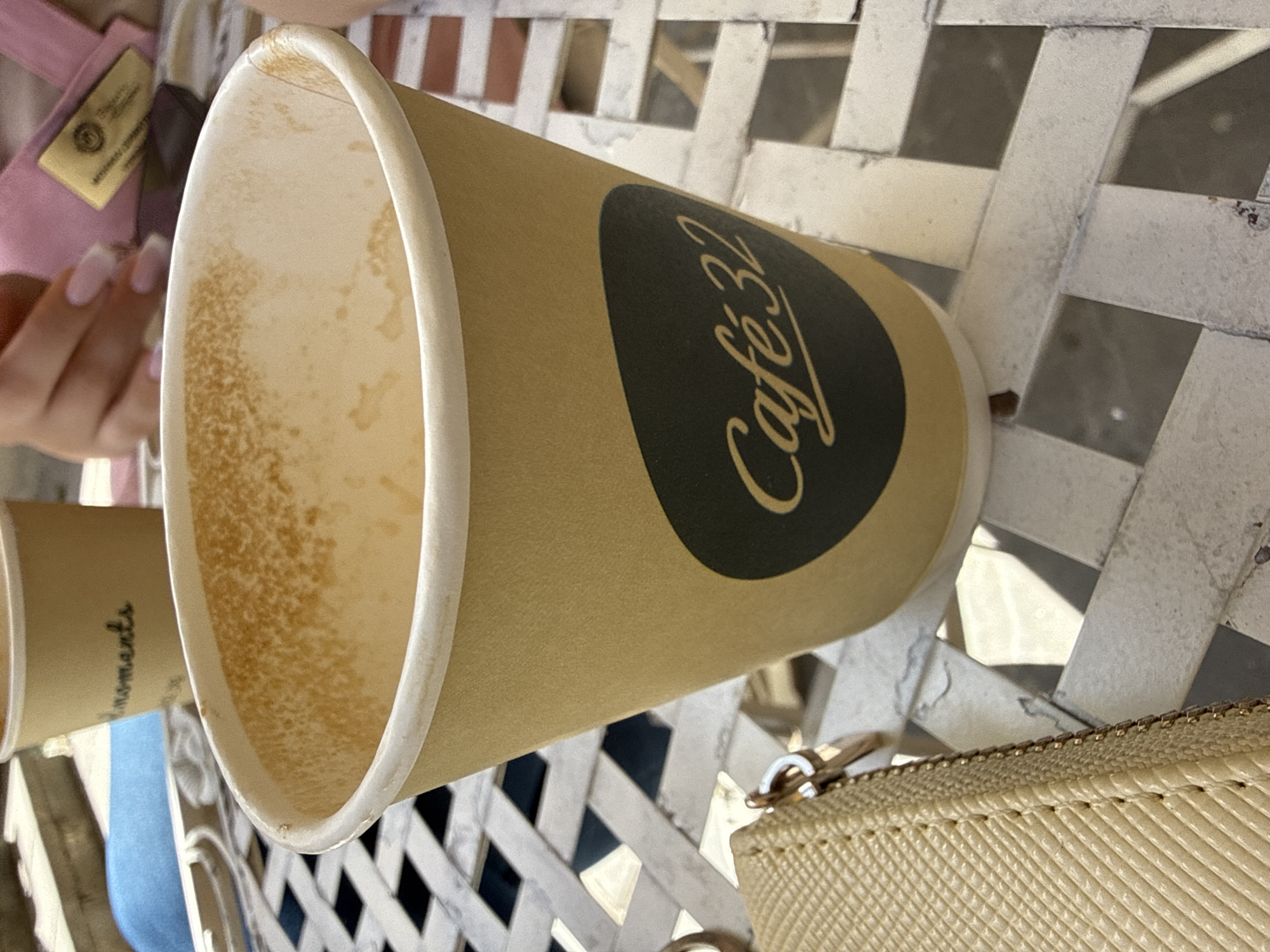 The image shows two partially empty paper coffee cups with the "Cafe 32" logo on a white metal latticed table, implying the setting is a café. The cups are sitting on the table amidst other items: a beige purse, a pink strap, and a hand with manicured fingernails. The location is likely Sofia, Bulgaria, based on the café name. The environment suggests an outdoor or semi-outdoor seating area due to the patterned table and latticed structure. The presence of used coffee cups indicates recent or ongoing consumption, with a person possibly sitting at the table. The overall scene depicts a casual, social setting at a café.