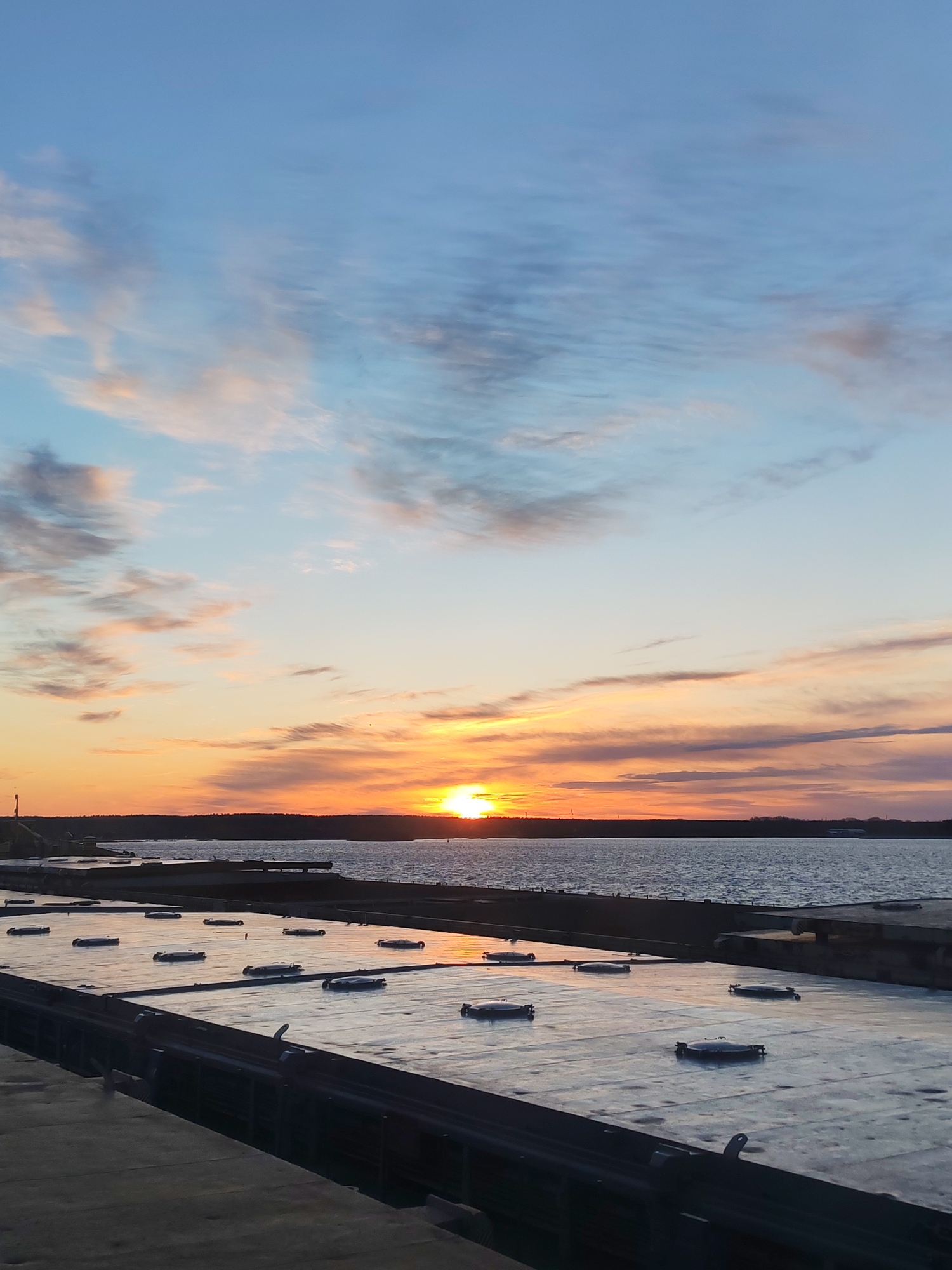A landscape photograph captures a sunset scene in an indeterminate location. The horizon is divided into three distinct segments. The upper portion of the image displays a sky with blue and light-colored clouds, transitioning to an orange and pink hue as it approaches the horizon where the sun is setting.
Below the sky, a body of water is visible, extending from the horizon to the middle ground of the photograph. The surface of the water reflects the colors of the sunset, enhancing the overall visual impact.
In the foreground, a dark, possibly metallic surface, marked with rectangular sections and hatch-like objects, dominates the lower part of the image. These features suggest an industrial or transportation context.
The image as a whole provides a contrast between the natural beauty of the sunset and the man-made elements in the foreground.