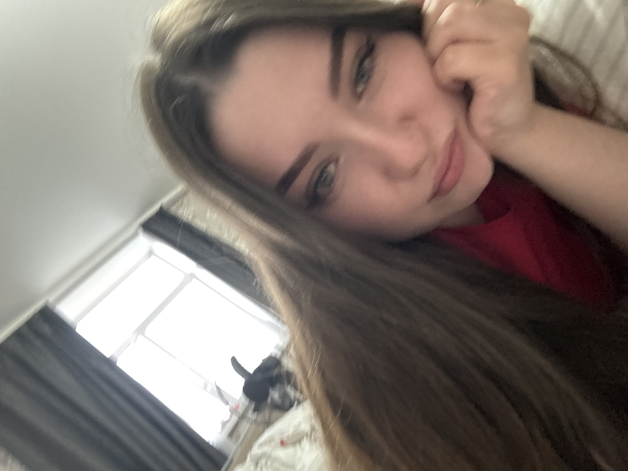 A slightly tilted, close-up indoor shot features a young woman in Savenki, Russia. She is the primary subject, looking towards the camera with a gentle, relaxed expression. Her long, straight light-brown hair frames her face, and her light-colored eyes are clearly visible. She wears a red top, and her hand is resting near her temple or cheek. In the blurry background, a window with bright light filtering through suggests it is daytime. Dark curtains partially cover the window. The setting appears to be a room, possibly a bedroom or living area. No text is visible in the image.