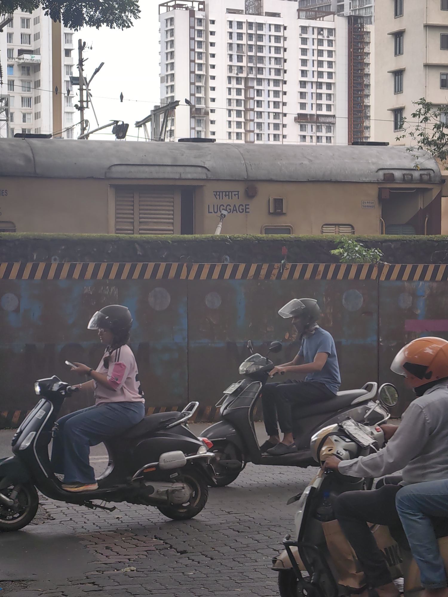 In Mumbai, India, on a street, three people ride scooters. The first scooter is black and is ridden by a woman wearing a pink shirt and blue jeans, with a black helmet. She is looking at her phone. The second scooter, also black, is ridden by a man in a blue t-shirt and black pants, wearing a black helmet. The third scooter is light beige and is ridden by a man wearing a grey shirt and blue jeans, with an orange helmet. Behind the scooters is a train car with the word "LUGGAGE" written in English and Hindi. The street has a cobblestone texture with some patches of water. In the background, there are tall buildings and trees.