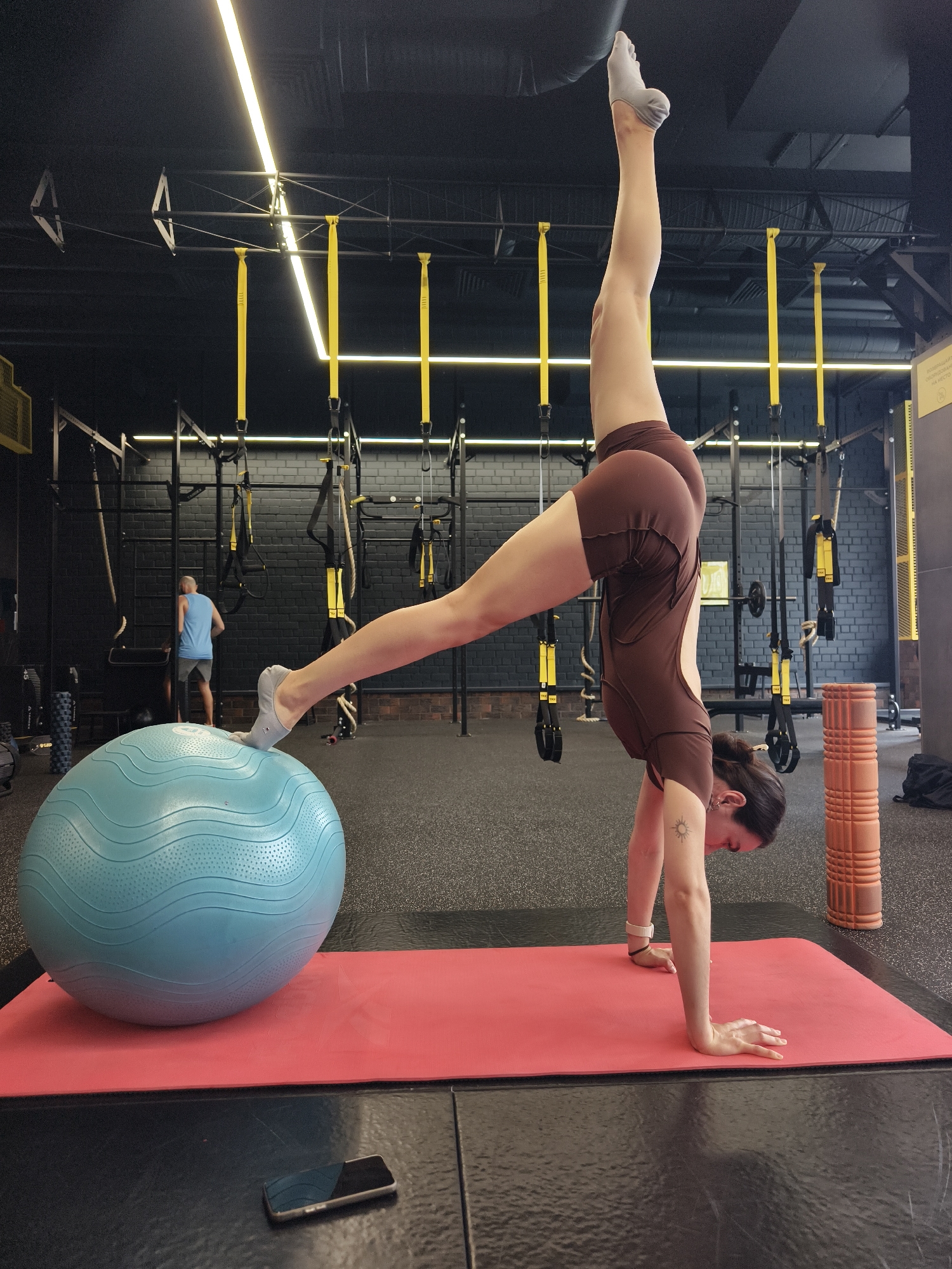 This image captures an indoor fitness facility, specifically a gym, in Tyumen, Russia. In the foreground, a female individual with dark hair tied back is performing a handstand on a red exercise mat. She is wearing a brown, form-fitting athletic outfit and grey grip socks. Her right leg is extended vertically upwards, while her left foot is placed on a large, light blue stability ball positioned on the mat. A small sun tattoo is visible on her left bicep, and she wears a light-colored wristband on her left wrist. A black smartphone is lying face down on the black floor tiles next to the mat.
In the mid-ground, to the right, an orange, textured foam roller stands upright. Further in the background, a male individual wearing a blue tank top and grey shorts is visible, facing away from the camera, near a black metal fitness rig.
The gym environment features dark, possibly painted brick walls and a dark ceiling with exposed structural elements. Bright, linear LED lights, some emitting a yellow hue, are mounted overhead. The floor consists of black rubberized or padded tiles, with a distinct lighter black stripe running across it beneath the exercise mat. Numerous yellow TRX suspension training straps, black metal rigs, ropes, and various other fitness equipment are present throughout the background. A yellow sign with Cyrillic text, including "ОБОРУДОВАНИЕ НА МЕСТО" (EQUIPMENT IN PLACE), is visible on the wall in the central background.