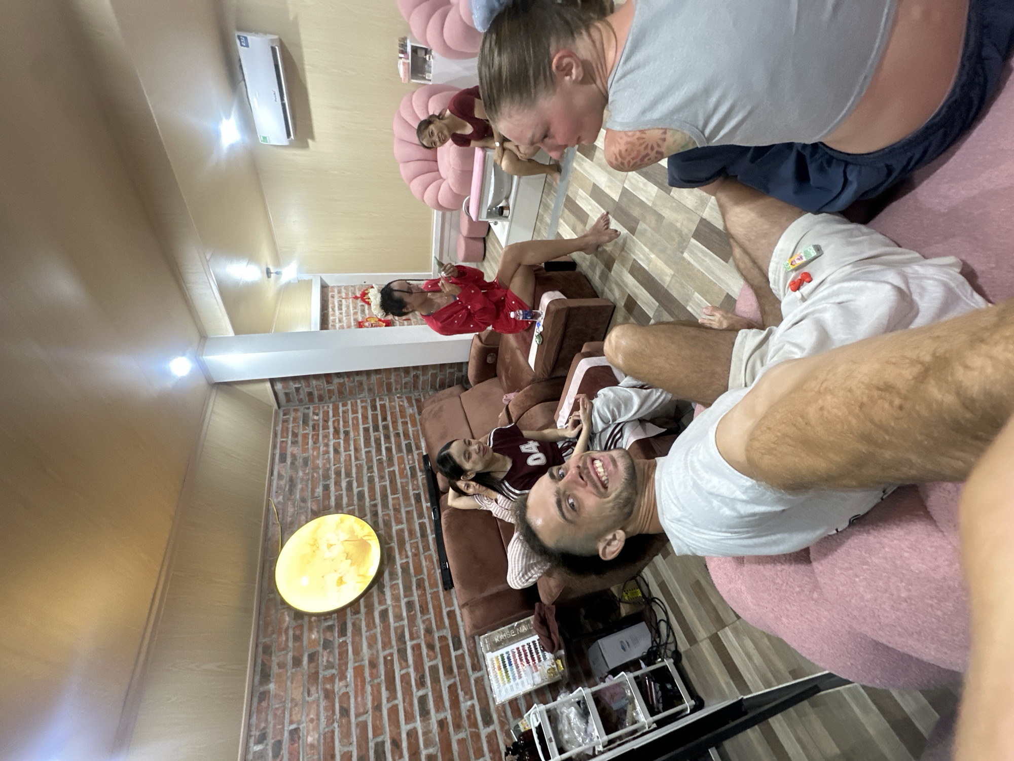 The image depicts an indoor scene, likely a nail salon, with several individuals present. A man in the foreground is seated on a pink beanbag chair, smiling at the camera. He is wearing a white t-shirt and light-colored shorts. To his right, a woman with blonde hair and tattoos on her arm is visible, wearing a grey tank top and dark pants. She is looking towards the man.
In the background, on a brown sofa, a young woman with dark hair is seated, wearing a maroon t-shirt with the number "04" on it and striped pants. Another woman is partially visible behind her. To the left of the sofa, a multi-tiered shelf unit contains bottles and what appears to be nail polish samples. A brick wall is visible behind the sofa, adorned with a circular yellow light fixture.
Further back, a woman in a red outfit is seated on a chair, holding a small object, possibly a phone or a receipt. She is barefoot. In the middle ground, another woman is seated on a pink chair, smiling. Adjacent to her is a white pedicure station with a basin. The flooring appears to be wood or laminate in a plank pattern. An air conditioning unit is mounted high on the wall in the background. The overall impression is a relaxed gathering, potentially a group of friends or clients at a salon. The location is identified as Nam Hoa Lư Ward, Vietnam.