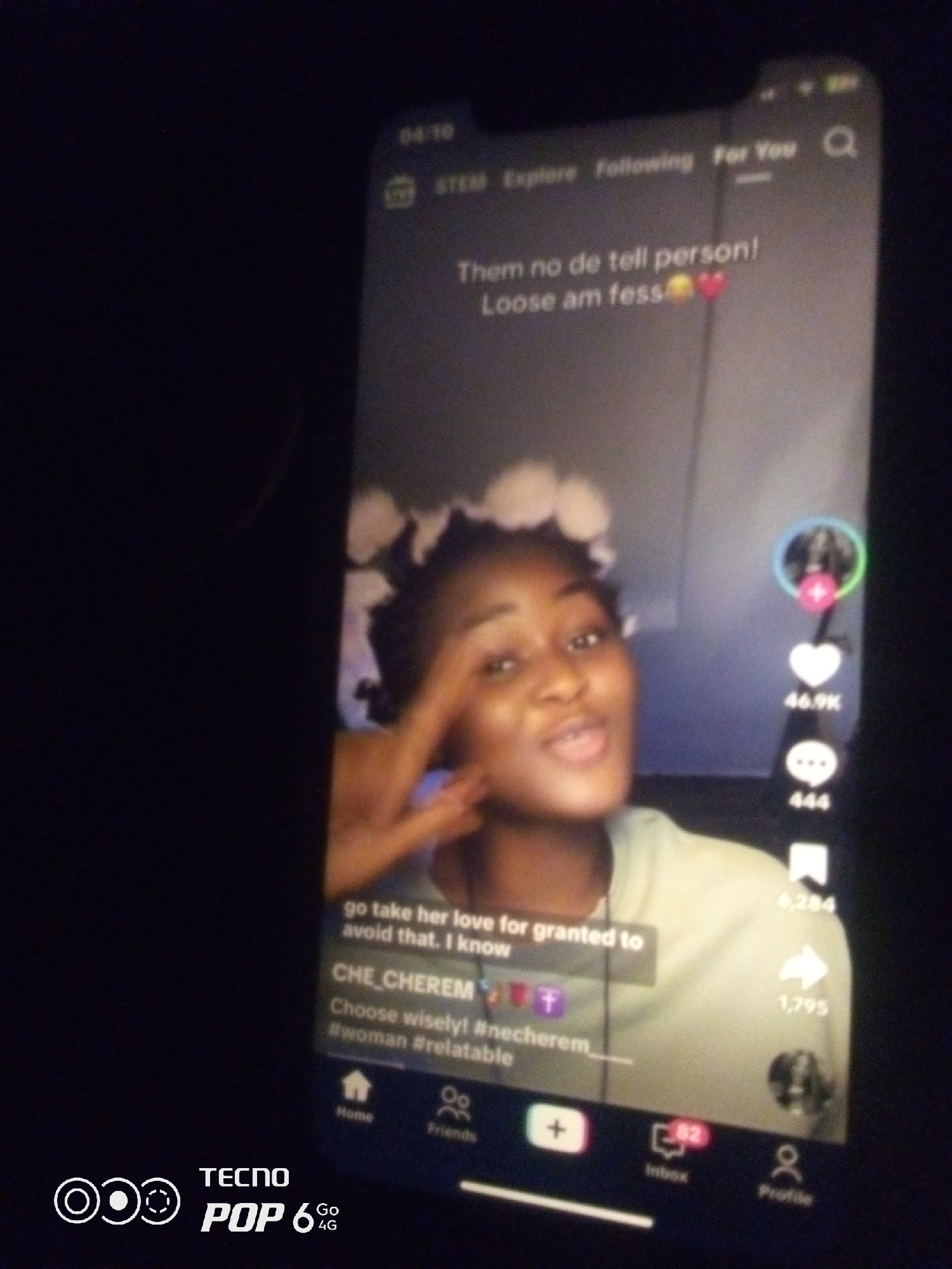 This is a close-up, low-light image of a smartphone screen displaying a social media video. The screen occupies most of the frame, with a dark background surrounding it. The video content features a young Black woman with flowers in her hair, smiling and speaking. Text overlays are visible on the screen, including "Them no de tell person! Loose am fess💛❤️", "go take her love for granted to avoid that. I know", "CHE_CHEREM", and "#woman #relatable". On the right side of the screen, there are social media interaction icons: a profile picture with a plus symbol, a heart icon with "46.9K" likes, a speech bubble icon with "444" comments, and a share icon with "1,795" shares. The bottom of the screen shows navigation icons for "Home", "Friends", a plus button for creating content, "Inbox" with "82" notifications, and "Profile". The phone's brand name "TECNO POP 6 Go 4G" is visible in the bottom left corner. The image does not provide any specific information or visual cues to determine its location within the city of Howayi, Nigeria.