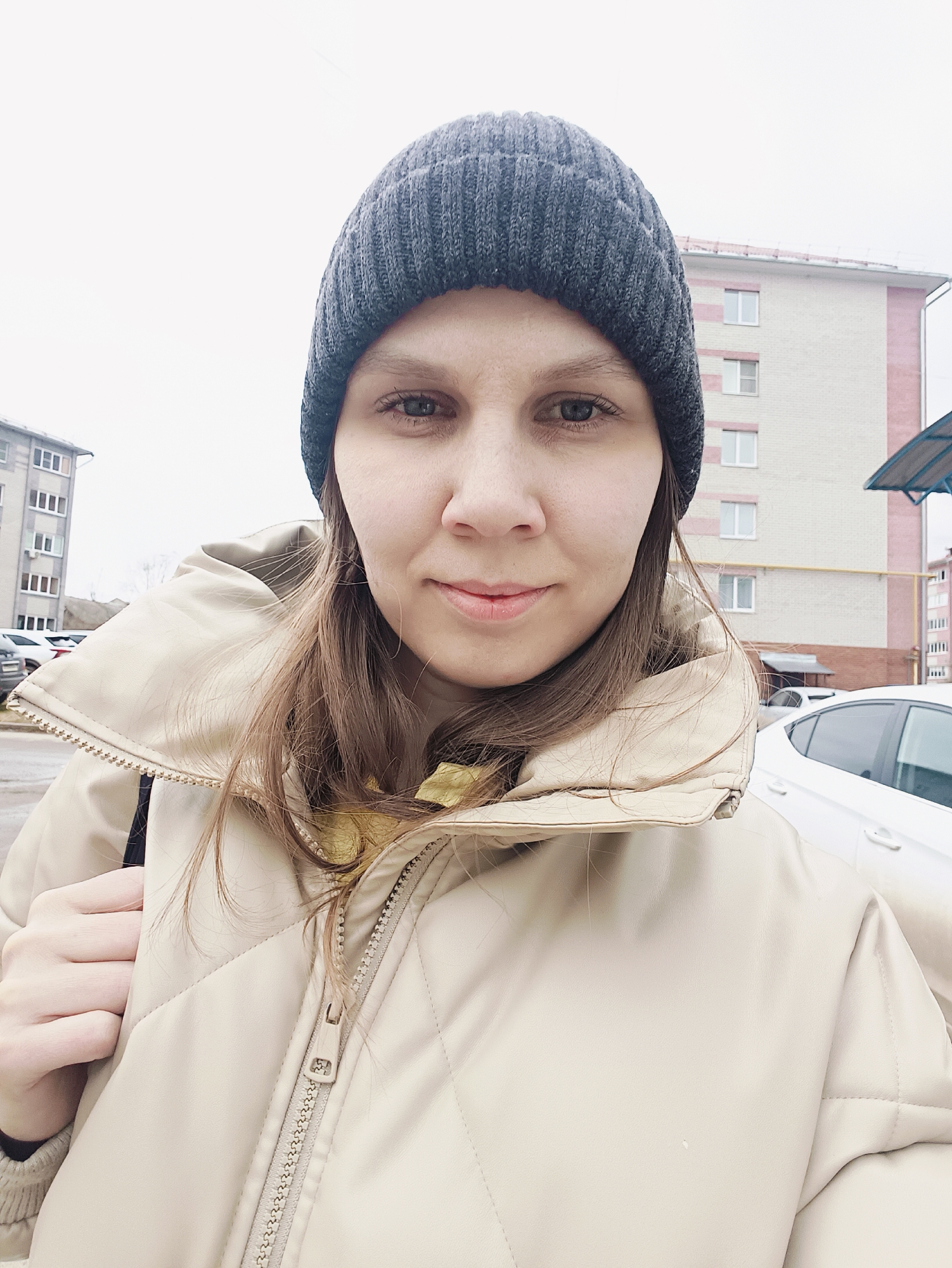 A person with a gray beanie and a beige jacket is taking a selfie outdoors in an urban environment. The individual's face is centered in the frame, displaying a neutral expression. Buildings are visible in the background, indicating a city setting. A white car is partially visible on the right side of the frame. The lighting suggests an overcast day.