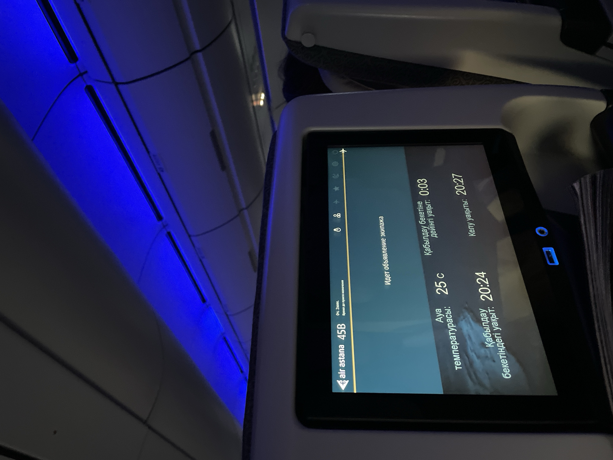 The image shows the interior of an airplane cabin, likely on an Air Astana flight, identified by the "air astana 45B" text on the screen. The scene is dimly lit with blue overhead lighting. A personal inflight entertainment screen is prominent in the foreground, displaying information in what appears to be a mix of Cyrillic and Latin alphabets. Text on the screen indicates "Идет объявление экипажа" (Crew announcement in progress), "Ауа температурасы: 25 C" (Air temperature: 25 C), "Қабылдау бекетінгі уақыт: 20:24" (Arrival station time: 20:24), "Қабылдау бекетіне дейінгі уақыт: 0:03" (Time until arrival station: 0:03), and "Келу уақыты: 20:27" (Arrival time: 20:27). The screen also shows icons for various entertainment options and a small airplane symbol. Out of focus passengers are visible in the background, and airplane seatbacks are present. The image does not contain any information that would place the location in Beykoz, Turkey.