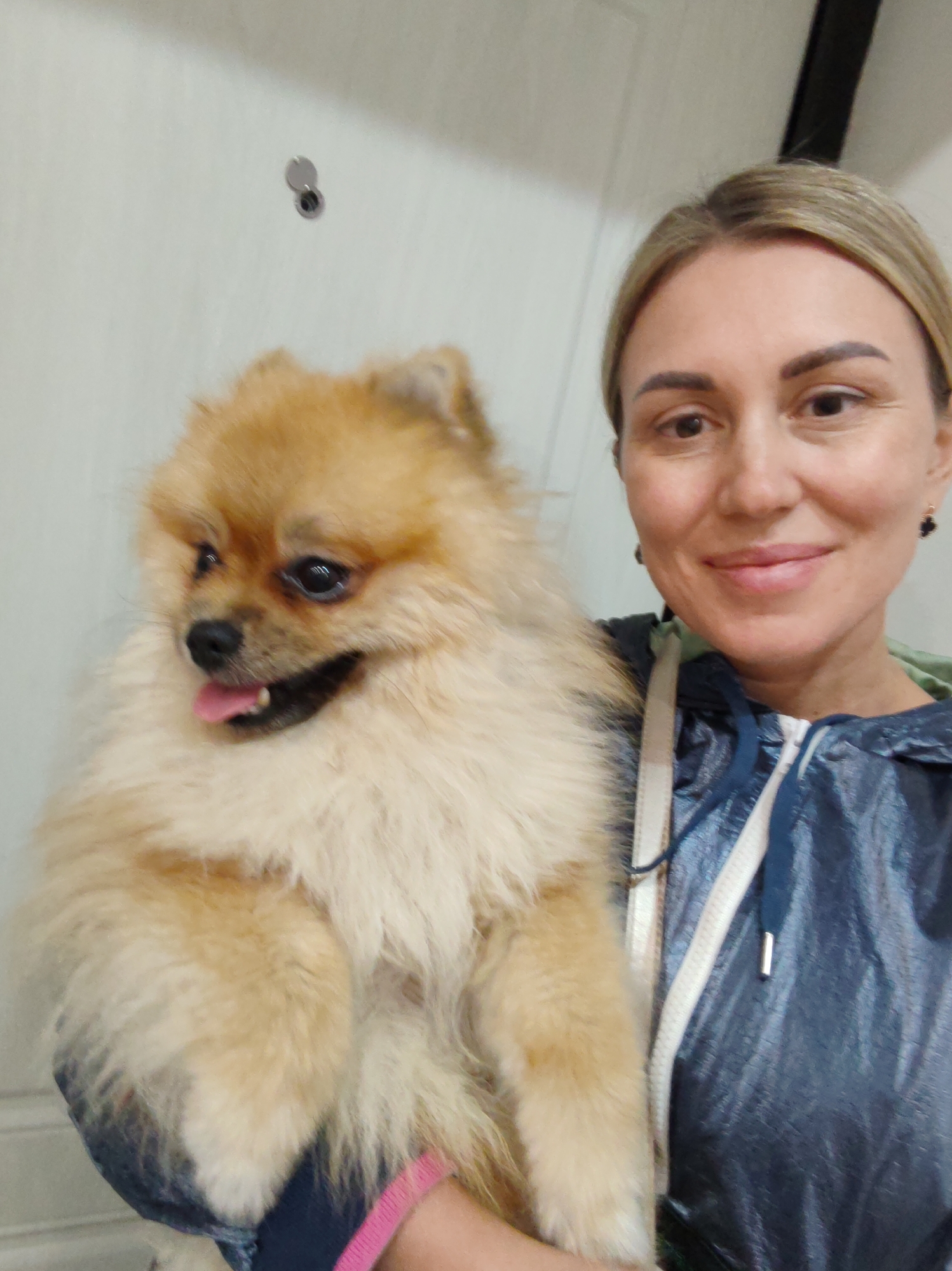 The image shows a medium shot of a woman holding a Pomeranian dog. The woman, who has blonde hair pulled back and is smiling, is wearing a blue, metallic-looking jacket over a green hoodie. The dog, a fluffy, light brown Pomeranian, is being held in her arms and appears happy, with its tongue sticking out. The background is a plain, light-colored wall with a door visible to the right. A small, circular object is attached to the wall in the upper left quadrant of the image, possibly a peephole or a decorative item. The lighting suggests it is daytime, and the indoor setting provides no clues about the weather. The overall mood is warm and affectionate, highlighting the bond between the woman and her pet. There is no visible text in the image.