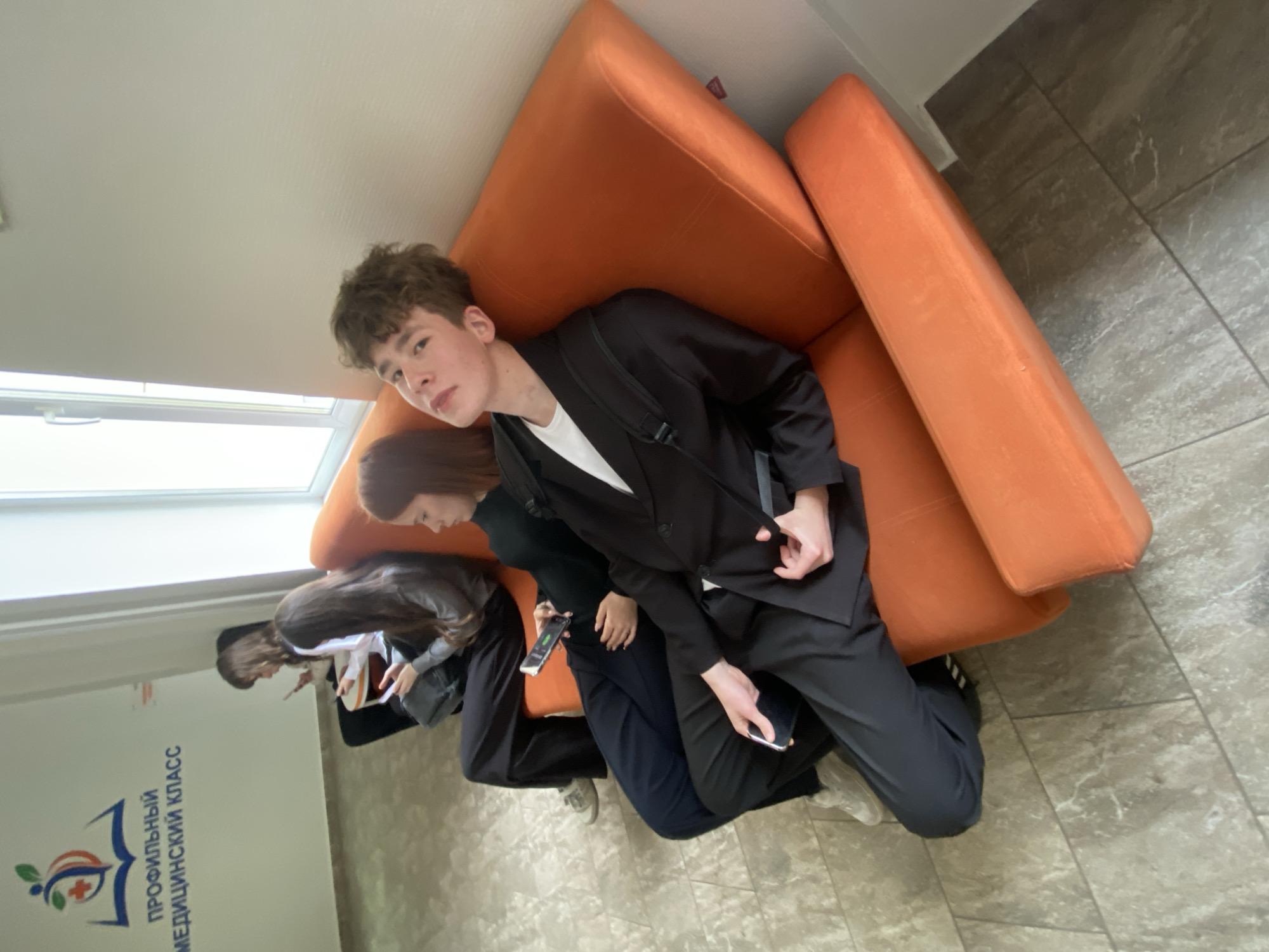 A high-angle shot shows a group of people seated on an orange couch in what appears to be a public space, potentially a school or medical facility, given the sign on the wall that reads "ПРОФИЛЬНЫЙ МЕДИЦИНСКИЙ КЛАСС" (Medical Class). The individuals are mostly young adults, possibly students, with one wearing a black suit jacket and holding a smartphone. Other individuals are also looking at their phones. The background includes a tiled floor, a window with natural light, and a glimpse of what might be more people in the distance.